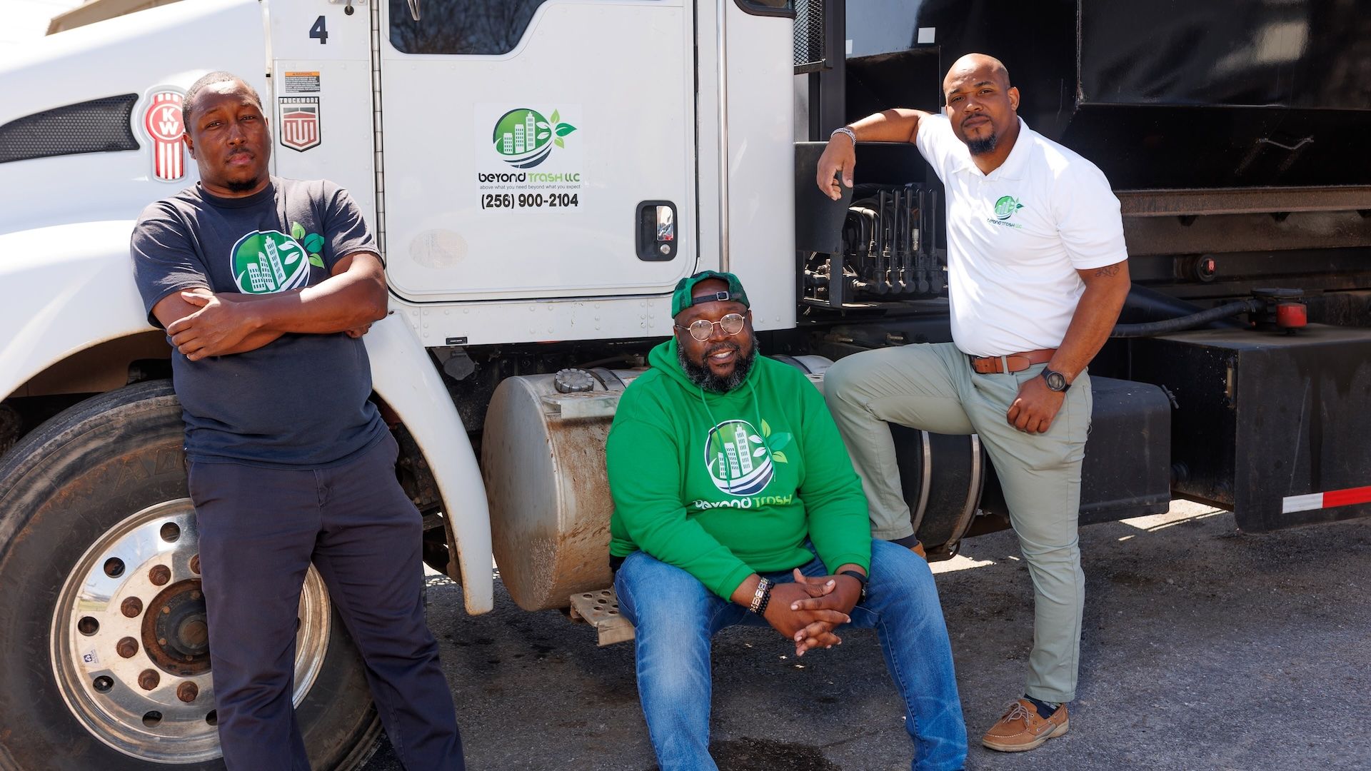 Three men pose beside a white semi-truck. Left in a dark T-shirt, middle in a green hoodie seated, right in a white polo. Logo on shirts reads Beyond Trash LLC with a cityscape and leaves.