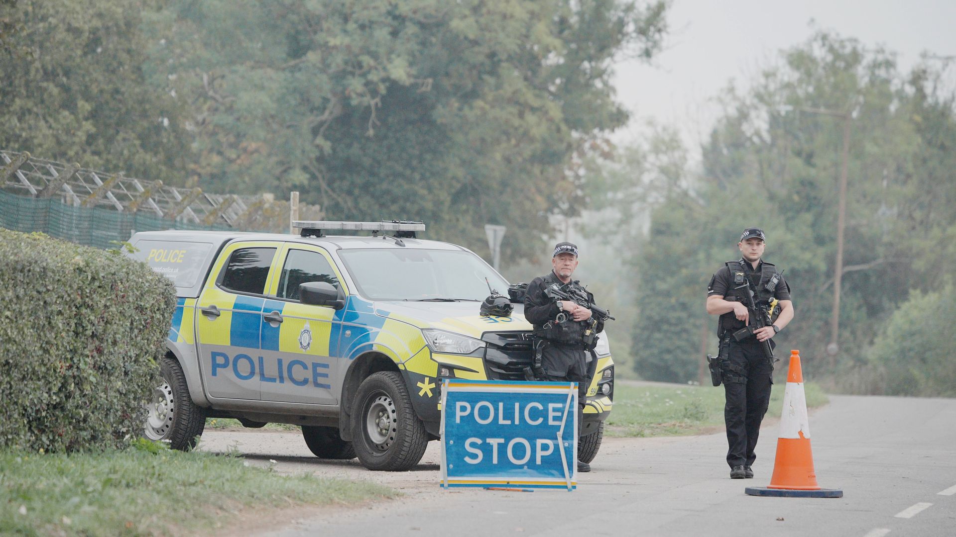 Two armed police officers in tactical gear stand beside a blue and yellow police SUV at a roadblock. A POLICE STOP sign and an orange cone mark the barrier, with trees in the background.
