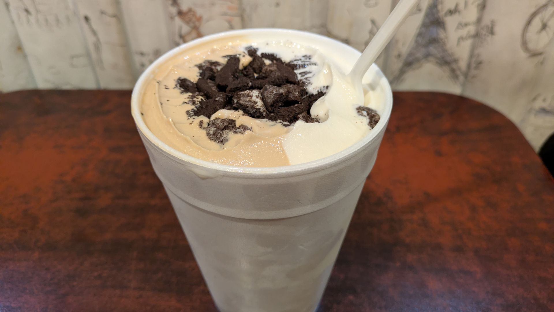Creamy dessert in a white foam cup topped with crushed chocolate cookies, divided into light brown and white halves, with a white plastic spoon inside. Background features a table and wall art.