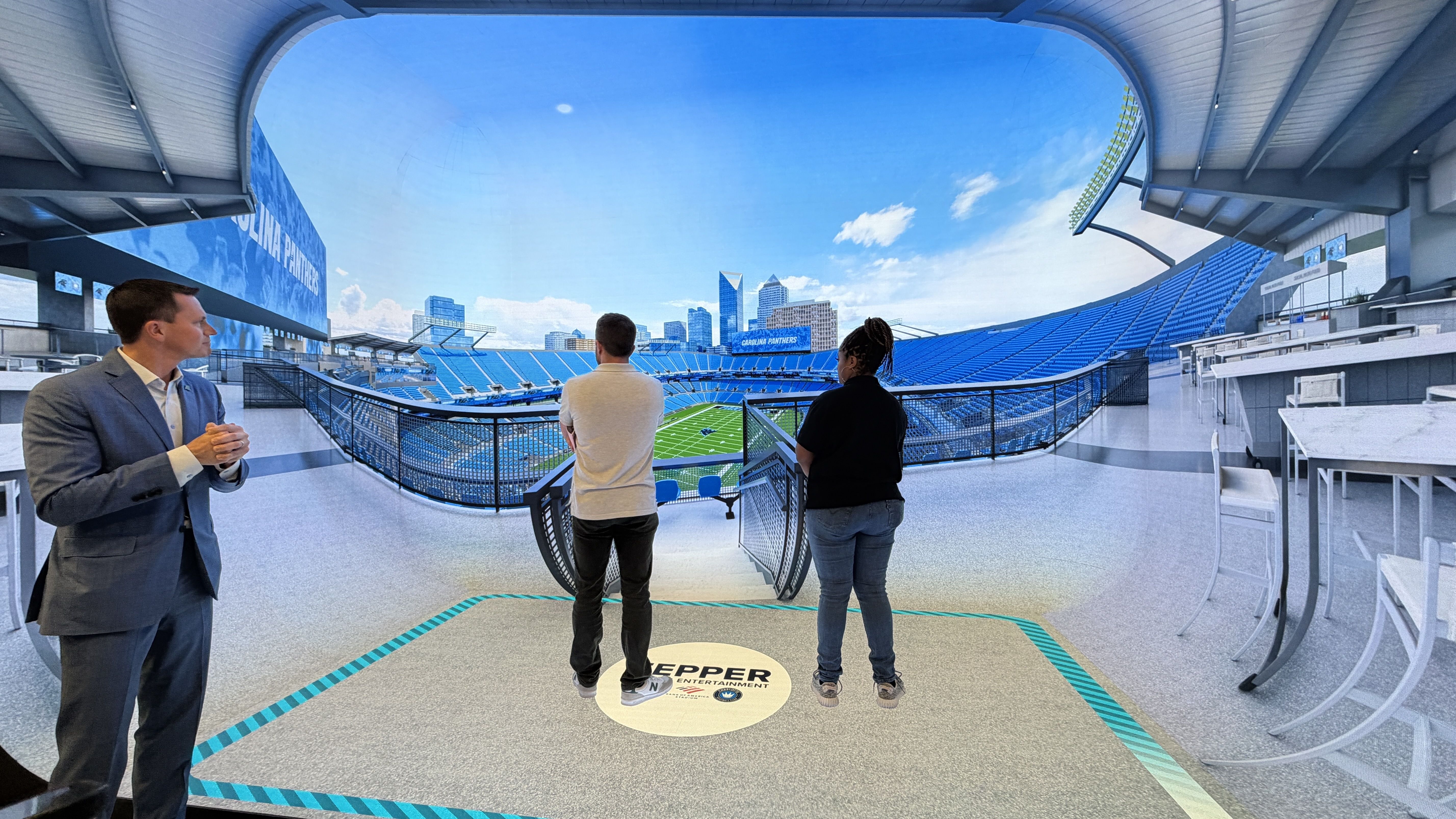 Indoor stadium viewing area with blue seats and curved ceiling. Three people stand at a railing facing the field: left a man in a gray suit, center a man in white shirt, right a woman in black.