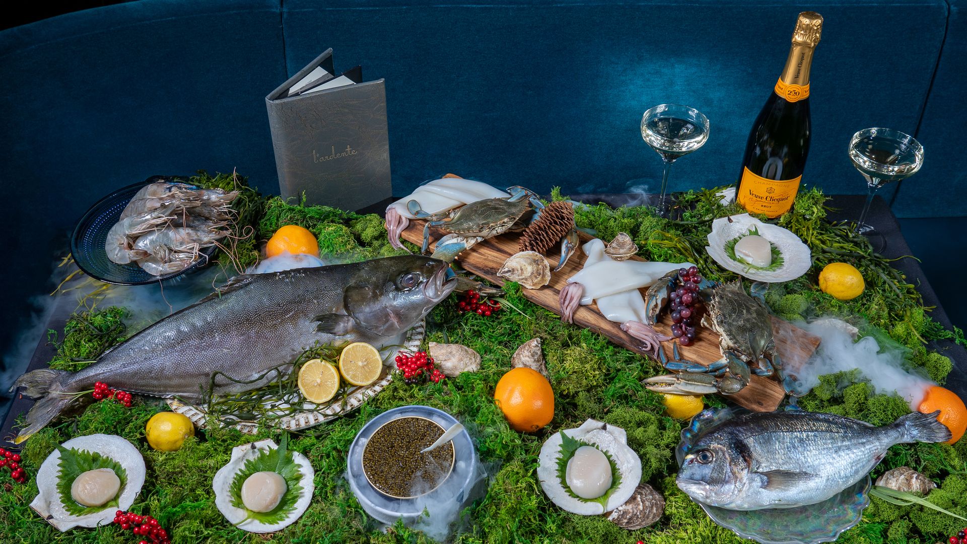 Luxurious seafood display with two whole fish, shrimp, squid, crab, scallops on shell, caviar, citrus fruits, champagne bottle and glasses on a bed of green moss and red berries.
