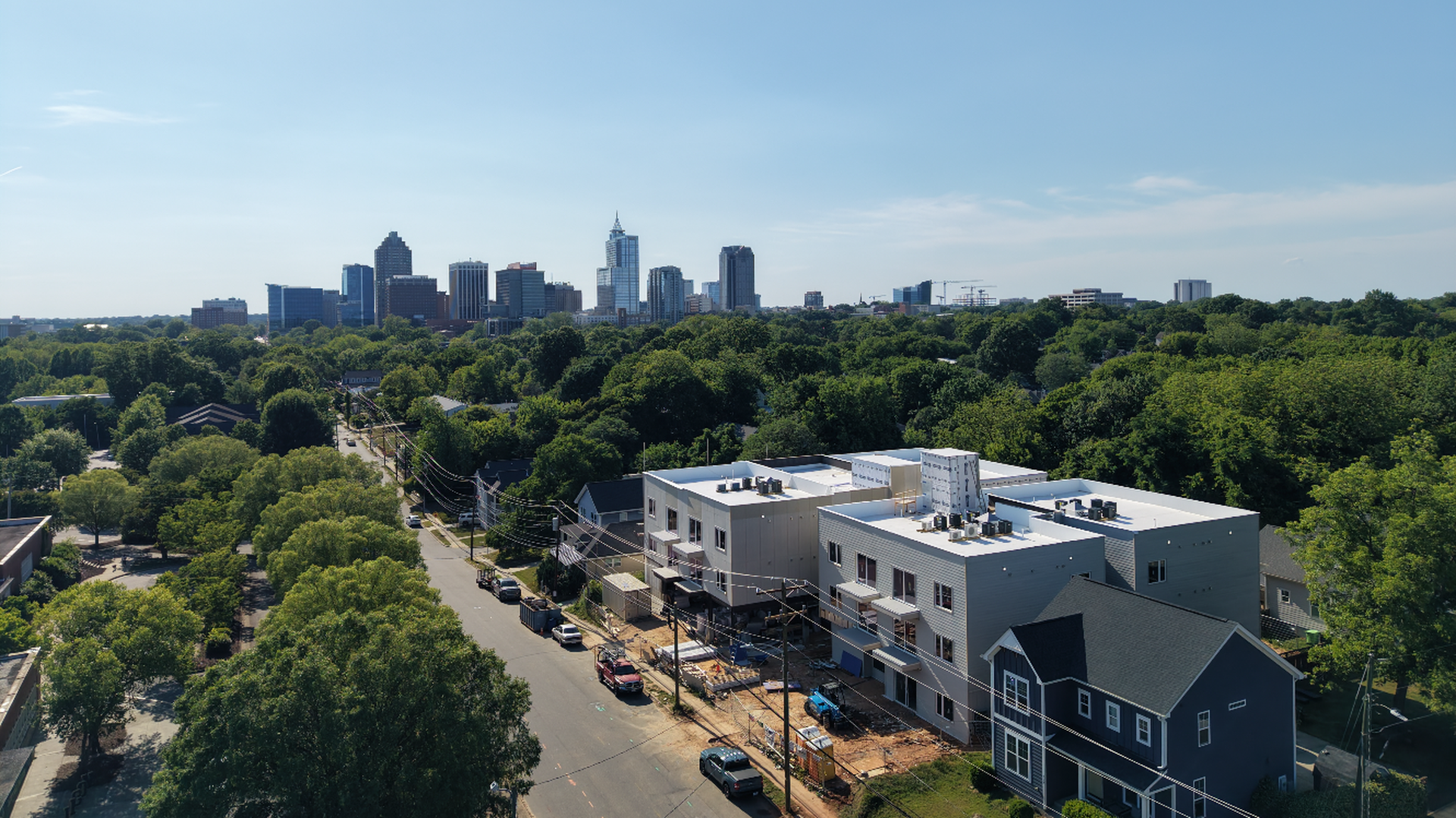 Aerial view of a city skyline on the horizon under a bright blue sky, with a tree-filled foreground and a cluster of low-rise buildings, including a white multi-story complex, along a street.