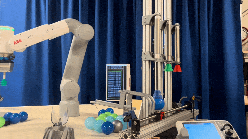 A blue plastic ball passes under a metallic assembly line as a robot arm moves to pick up another ball. A monitor flashes in the background in front of a blue curtain backdrop.