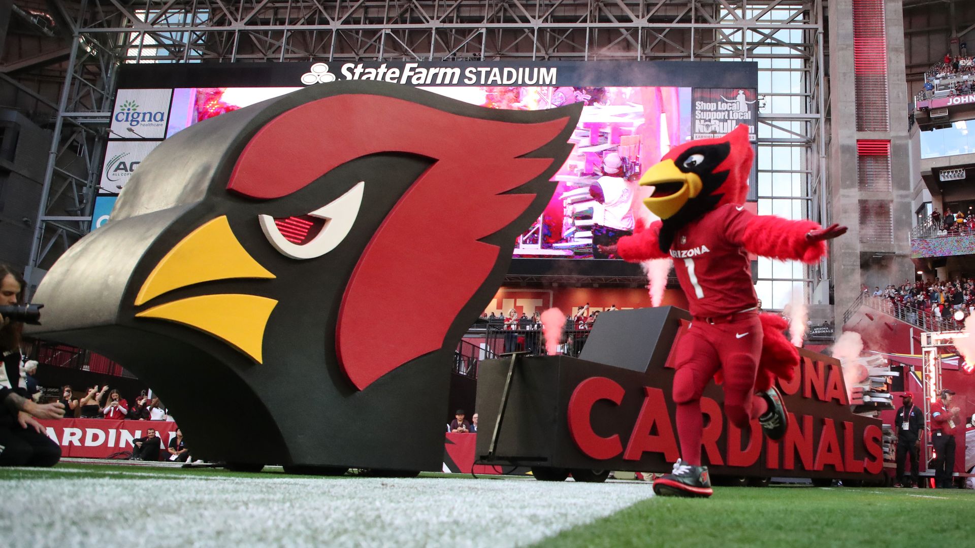 Arizona Cardinals mascot head on the field at State Farm Stadium, with a large video screen behind, metal trusses overhead, and cheering fans in the stands.