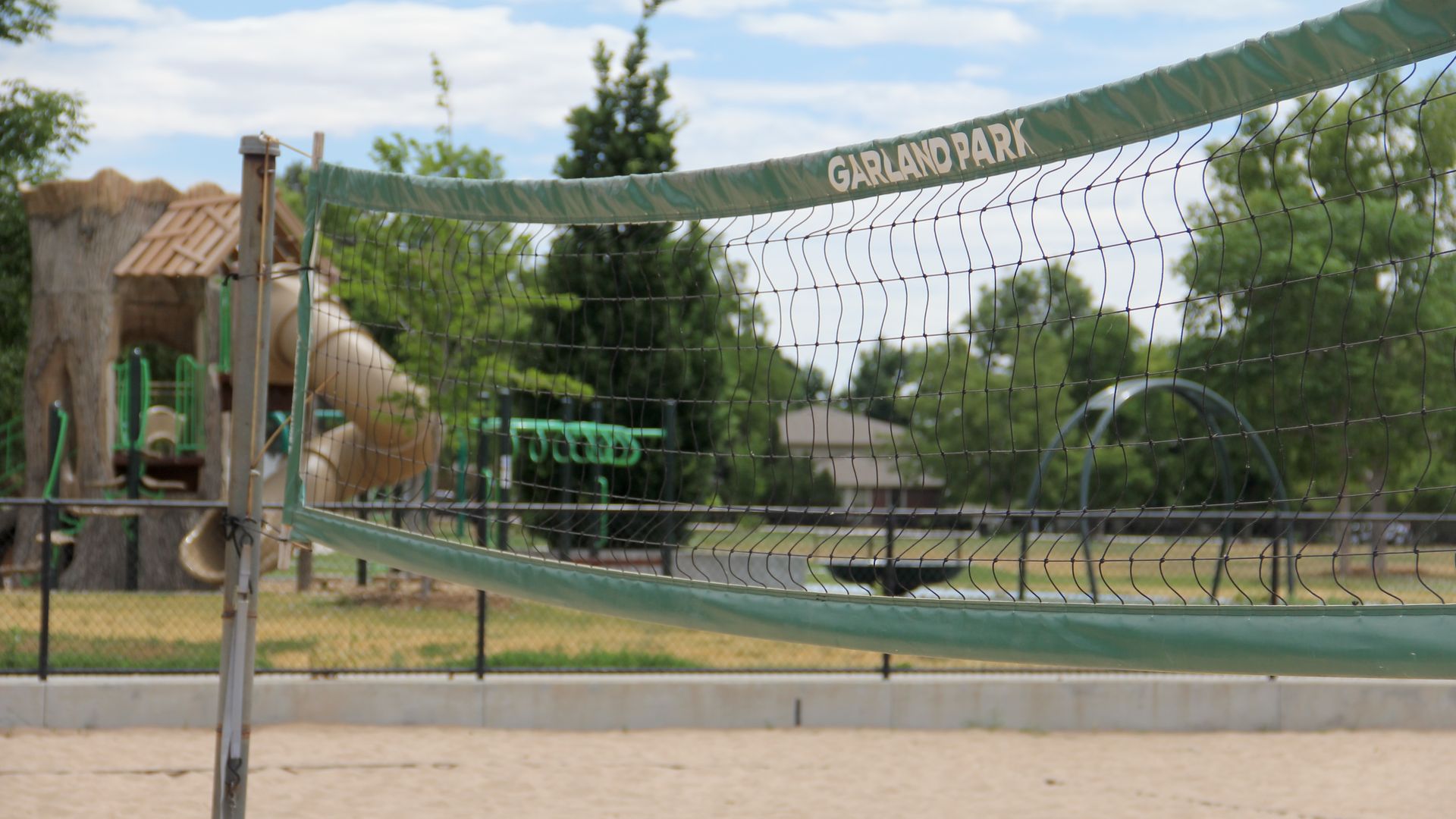 A green volleyball net during a sunny day. In the background, a children's playground is visible. 