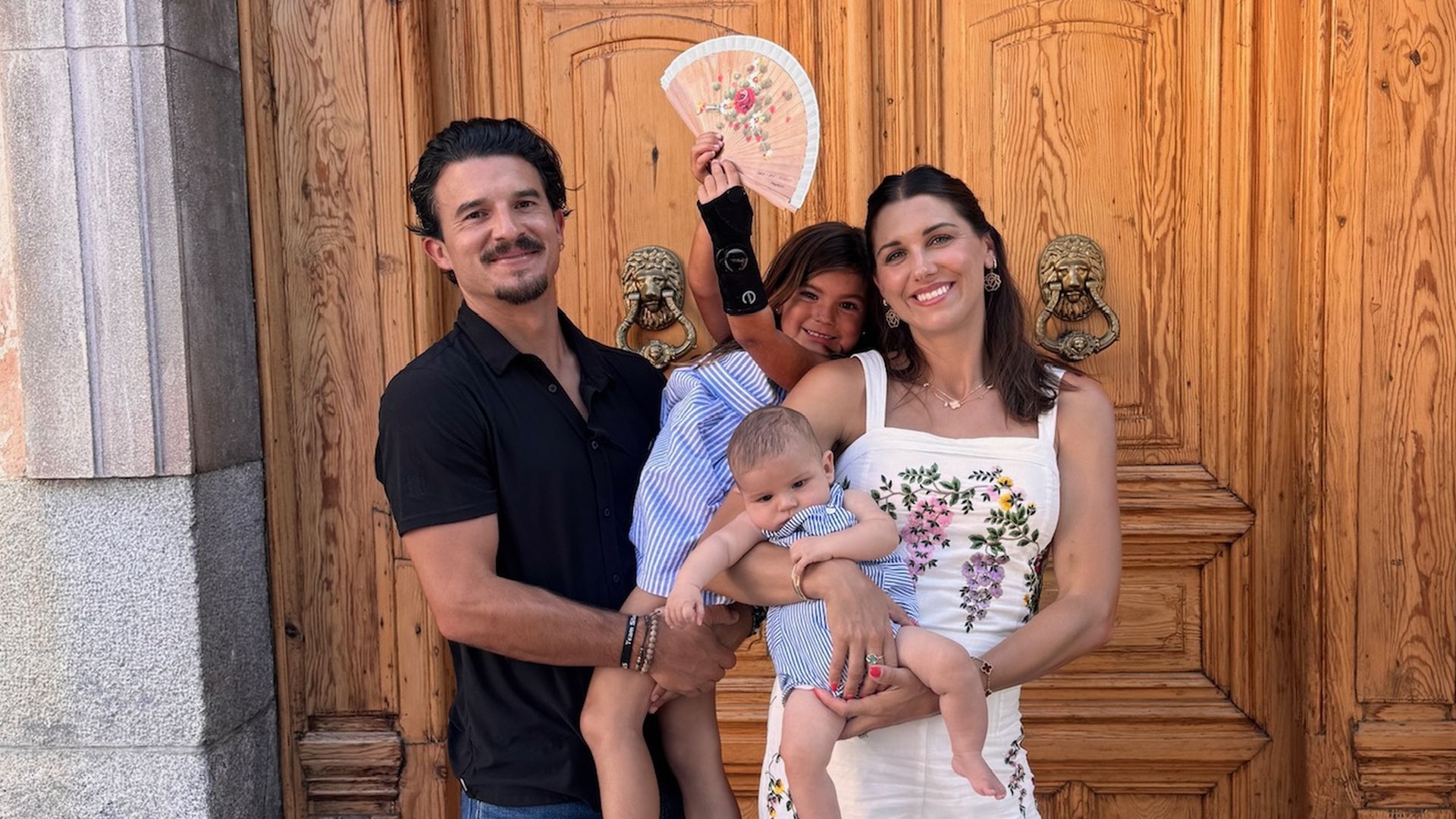 A family of four stands in front of a large wooden door with decorative knockers and the number 8 above. The man wears a black shirt and blue jeans. The woman wears a white floral jumpsuit, holding a baby. A young girl in a blue dress and white sandals holds a floral fan.