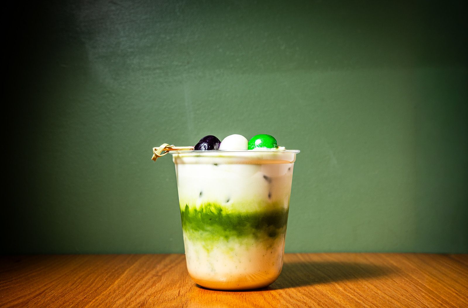 Layered dessert drink in a clear cup on a wooden table: white creamy top, green tea layer, pale bottom; topped with three round candies (black, white, green) on a skewer against a green wall.