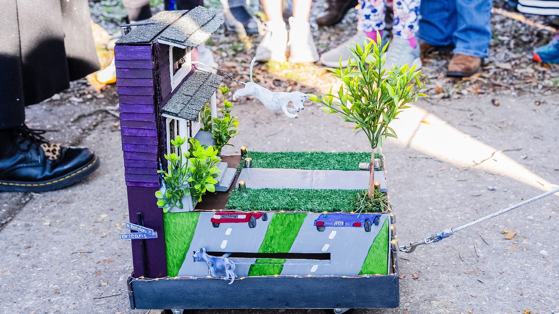 A shoebox float is pulled along by a strong. On the float, a tiny dog jumps out of a replica house window.