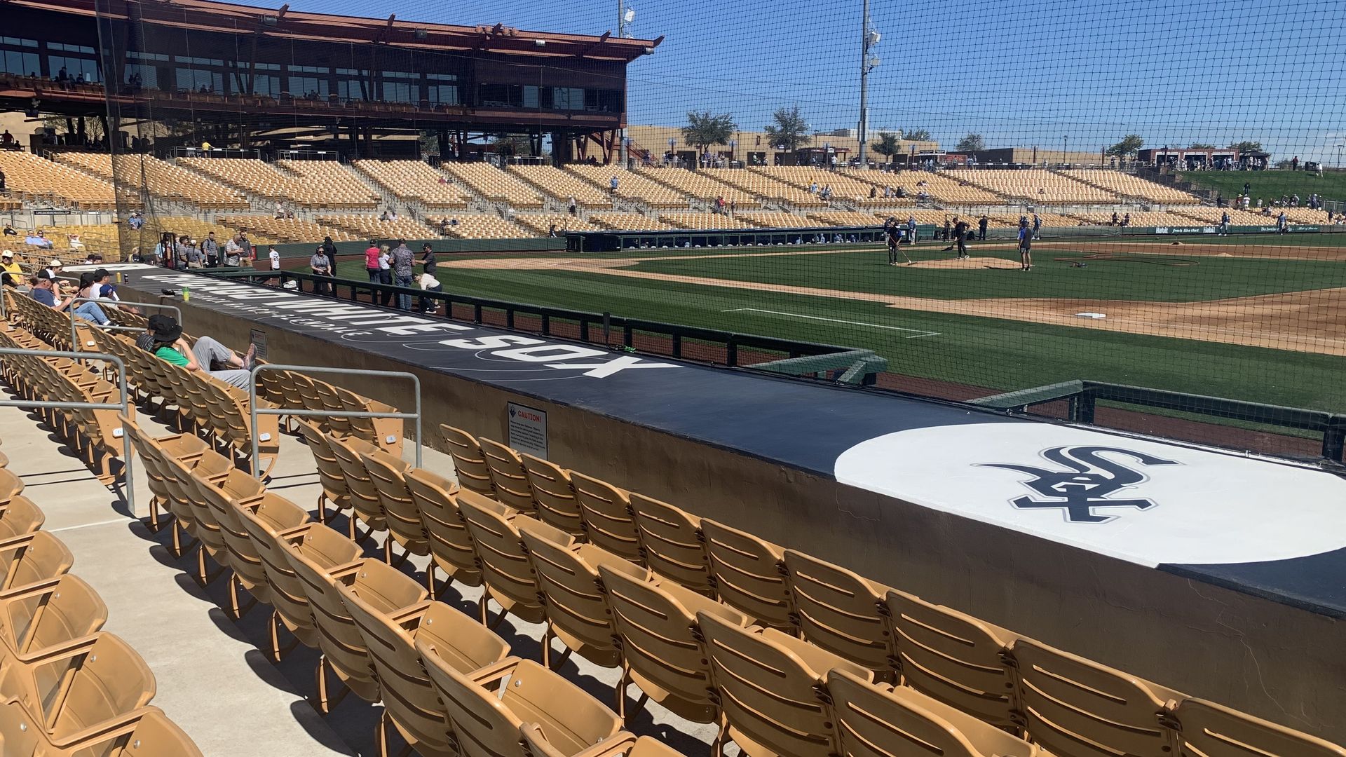 Empty tan seats at a baseball stadium with a green field and players on the field, blue sky, and a building in the background. The Chicago White Sox logo is visible near the seats.