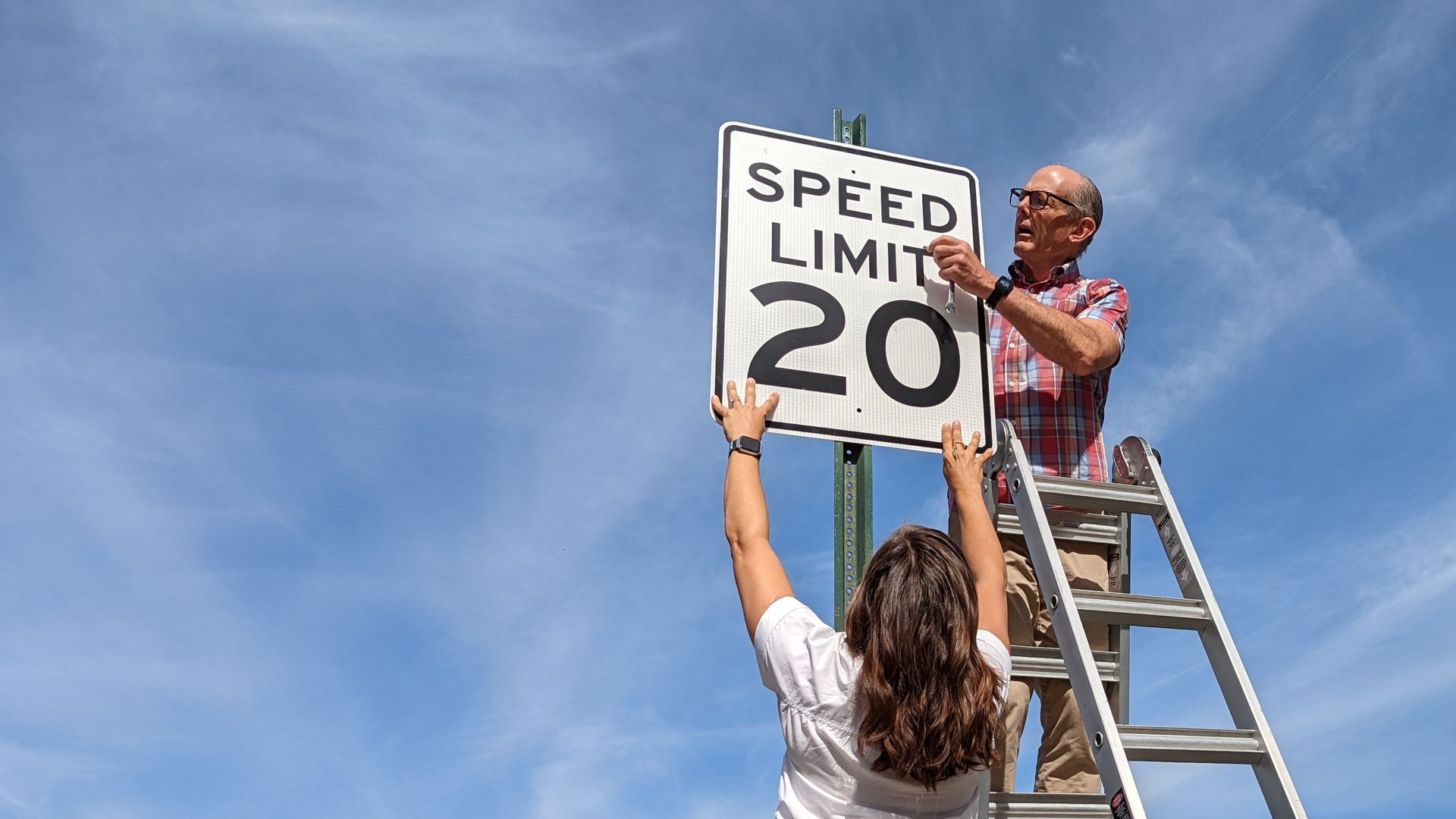 A woman and a man install a 20 mph sign on a signpost.