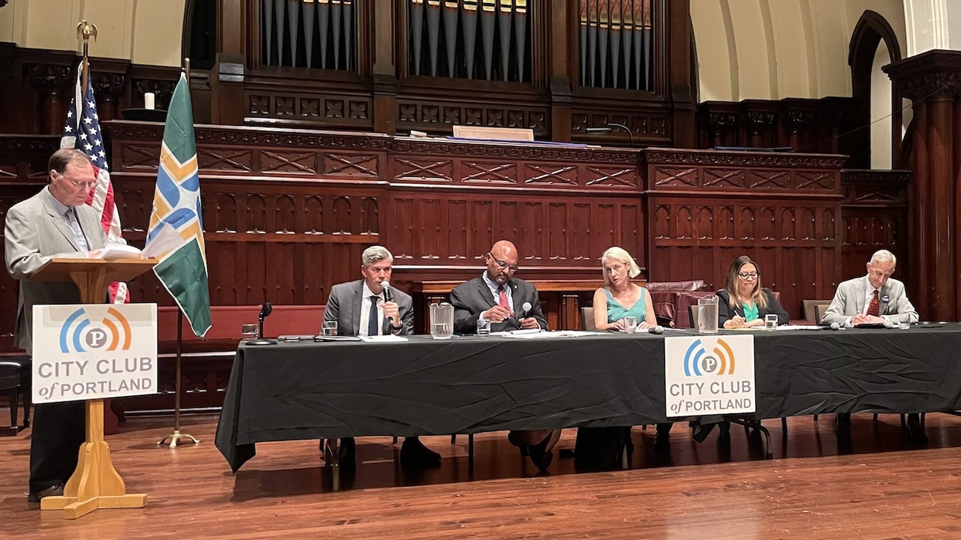 A photo of one person standing at a lectern and five others, seated behind a table with a black cloth.