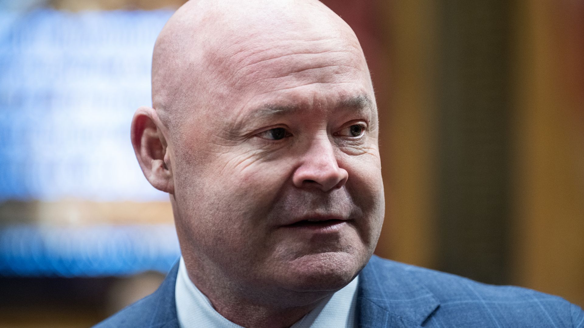 Close-up of a person man in a blue suit and striped tie, looking to the right. A gold badge is pinned to his lapel, with a blurred blue and brown background.