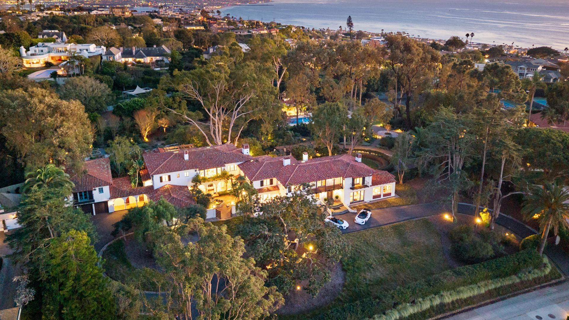 Aerial view of a coastal hillside neighborhood at sunset featuring a large red-tiled villa, lush trees, winding driveway, and the ocean in the distance.