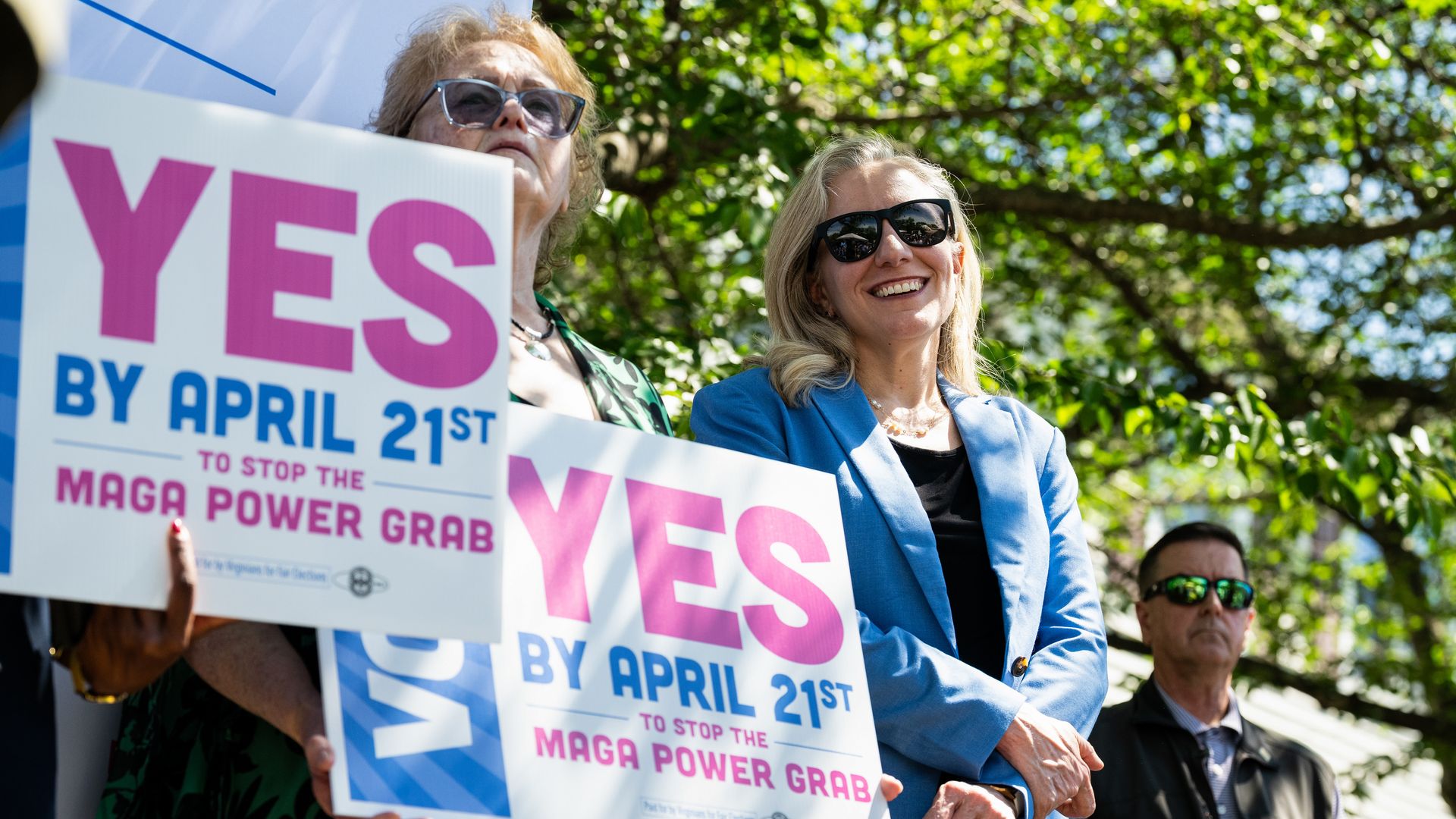 Virginia Gov. Abigail Spanberger, a a blue jacket, is shown with signs that say "YES By April 21st" 