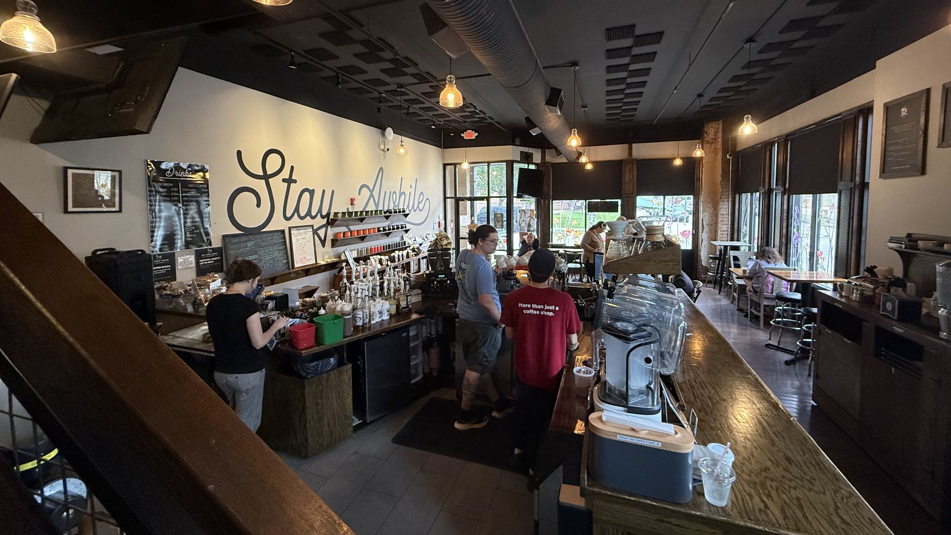 Cozy cafe interior with warm pendant lights and a long wooden bar. Customers queue at the counter; a cursive wall sign reads "Stay Awhile," and seating is visible by the windows in back.