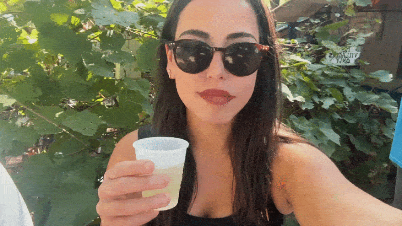 A woman with long dark hair, oversized black sunglasses, and red lipstick holds a small plastic cup of a light-colored drink outdoors, with green vines in the background.