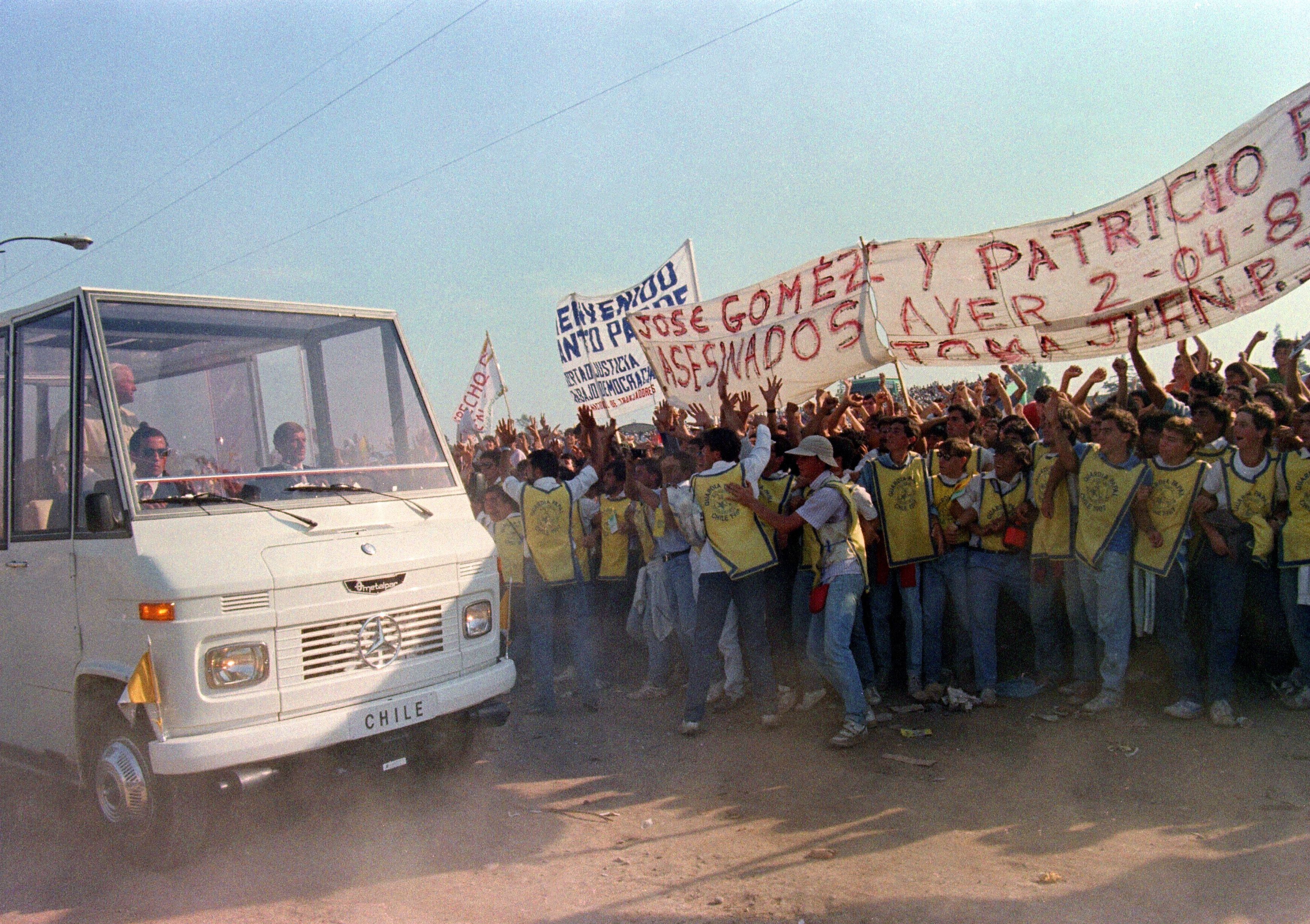 a crowd of protesters in yellow vest, on the right, hold up large signs as Pope John Paul II drives by in the Popemobile