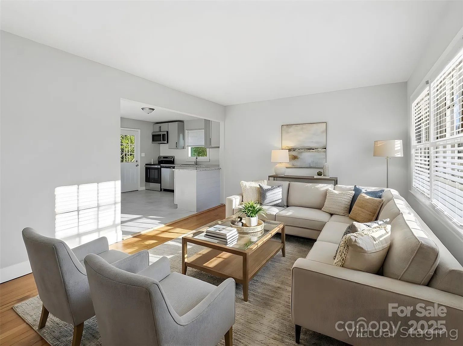 Bright living room with beige sectional sofa, two gray armchairs, wooden coffee table with decor, large windows with white blinds, and view into white kitchen.