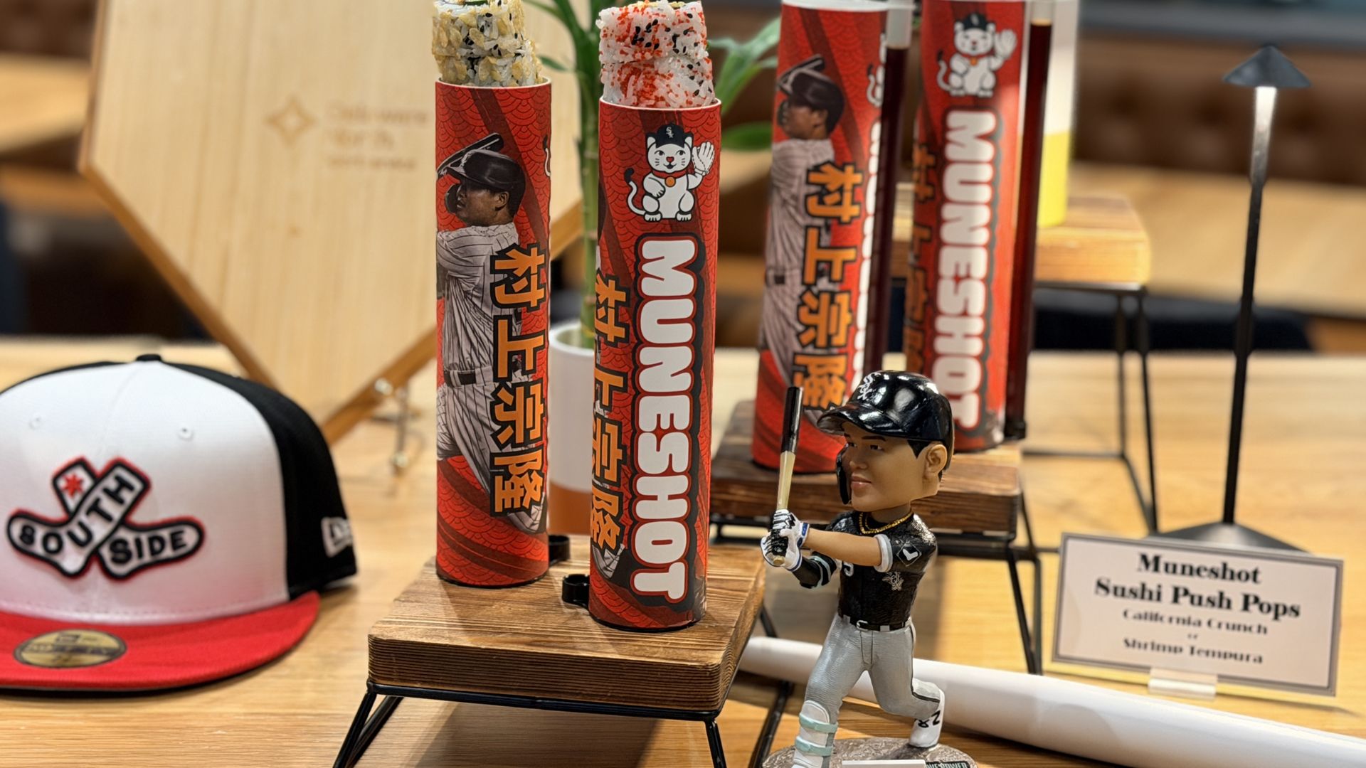 Sushi pops in red wrappers with cartoon cats stand upright on wooden bases; foreground includes a white and black cap and a toy figure, with a blurred shop interior in the background.