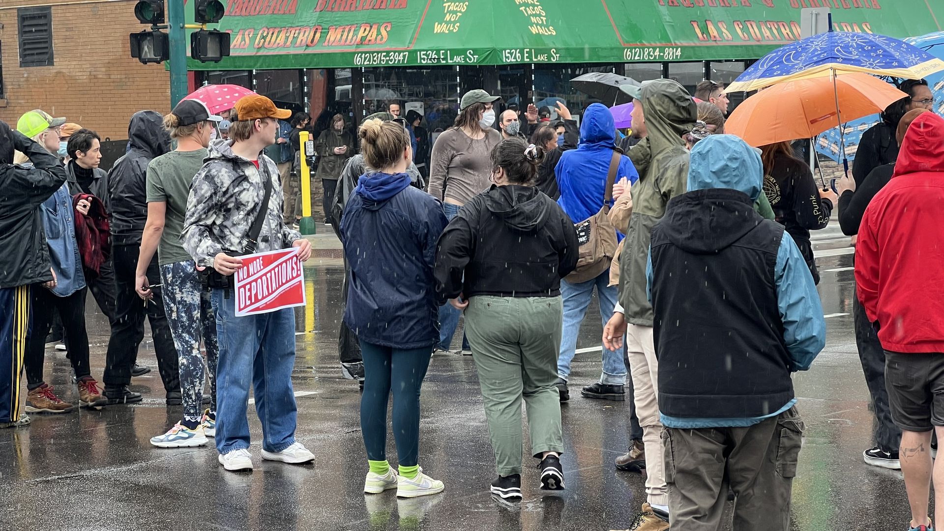 Protestors including one holding a sign saying "No More Deportations" stand in the middle of a street in front of a storefront with a green awning and the name "Taqueria y Birrieria Las Cuatro Milpas"