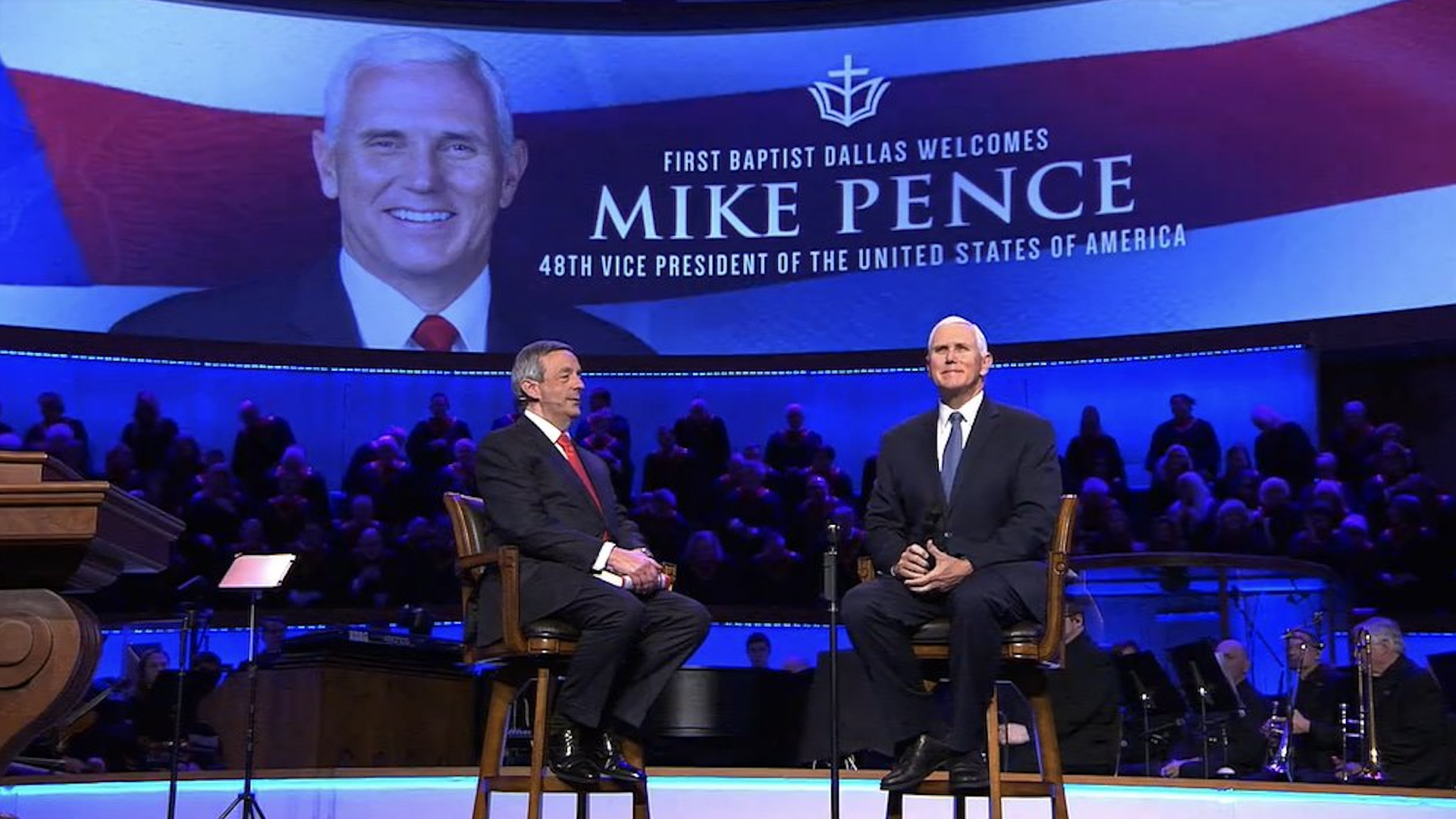 Two men in dark suits sit on a stage. Pence smiles like a happy school boy.