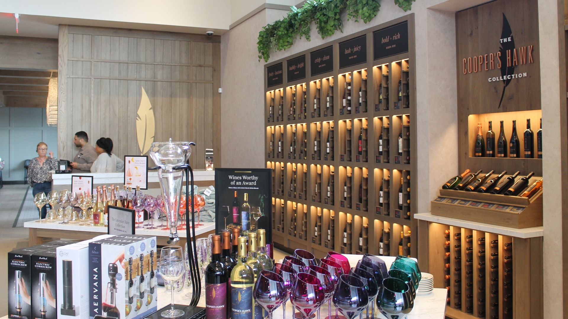 Interior of a wine store featuring wooden shelves with illuminated wine bottles, a display of colorful wine glasses, and a sign for The Cooper's Hawk Collection on the wall.