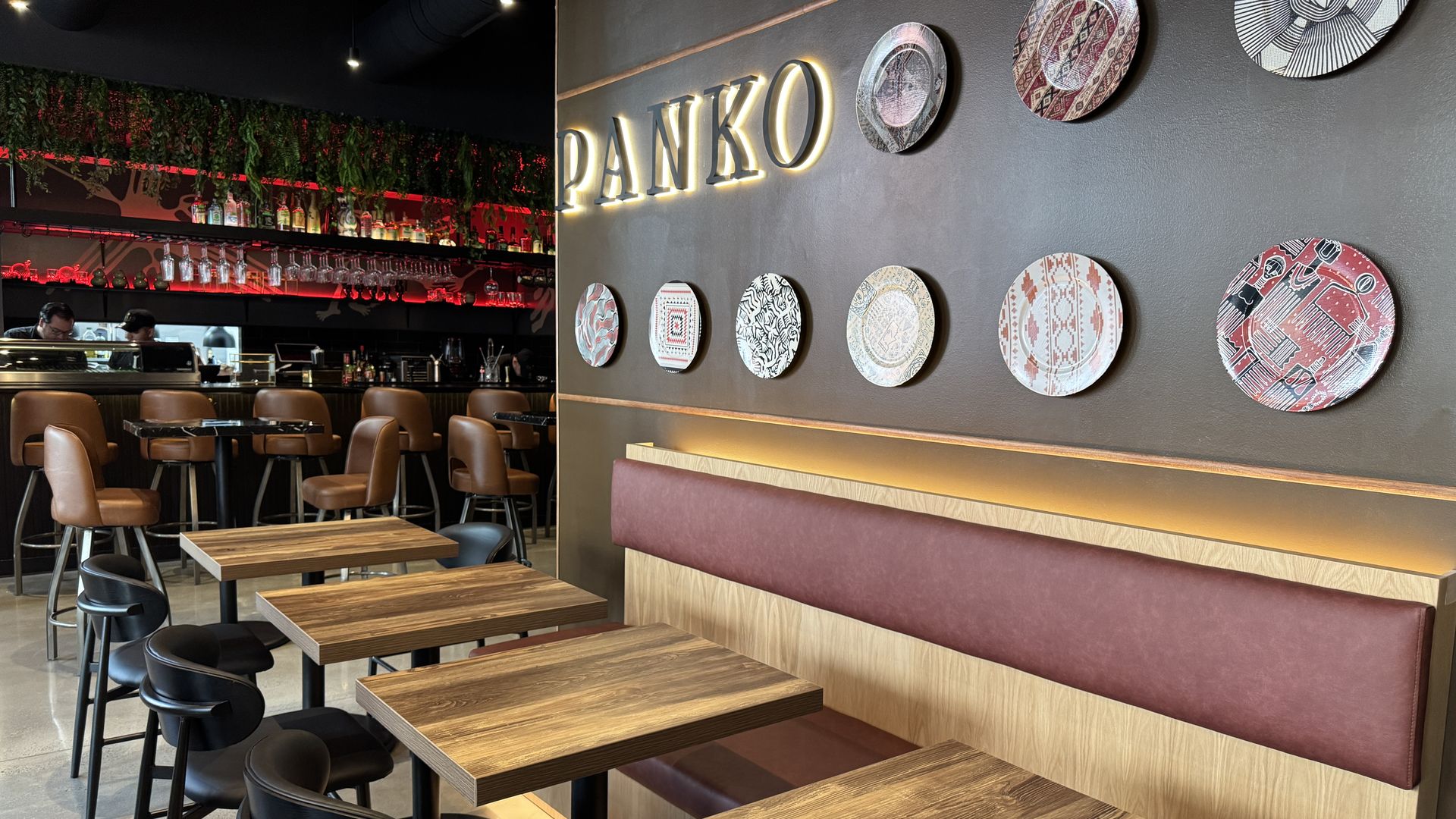 Interior of a modern restaurant named PANKO with wooden tables, black and brown chairs, red cushioned bench, decorative patterned plates on a dark wall, and a bar with hanging glasses and red lighting.