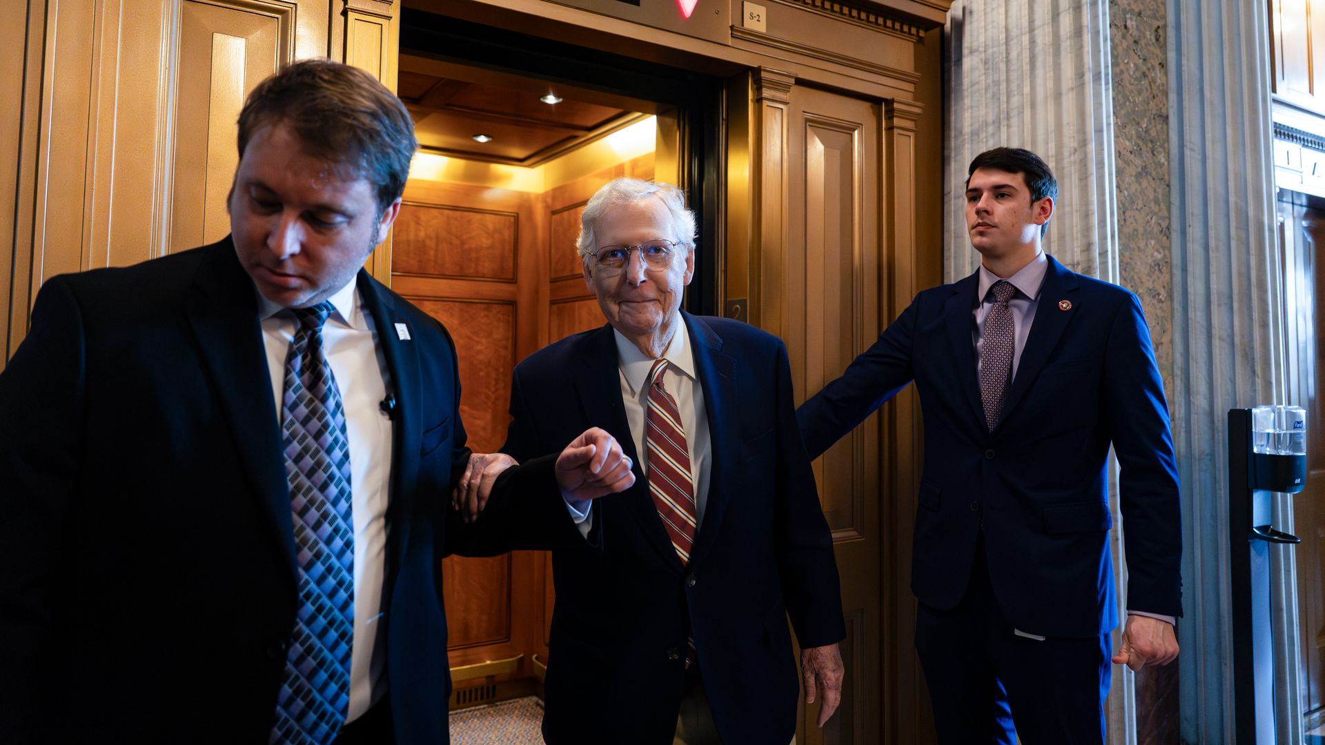 Three men in suits near an open elevator with wood paneling; the middle man with white hair and glasses smiles, flanked by two men, one holding his arm, in a formal setting.