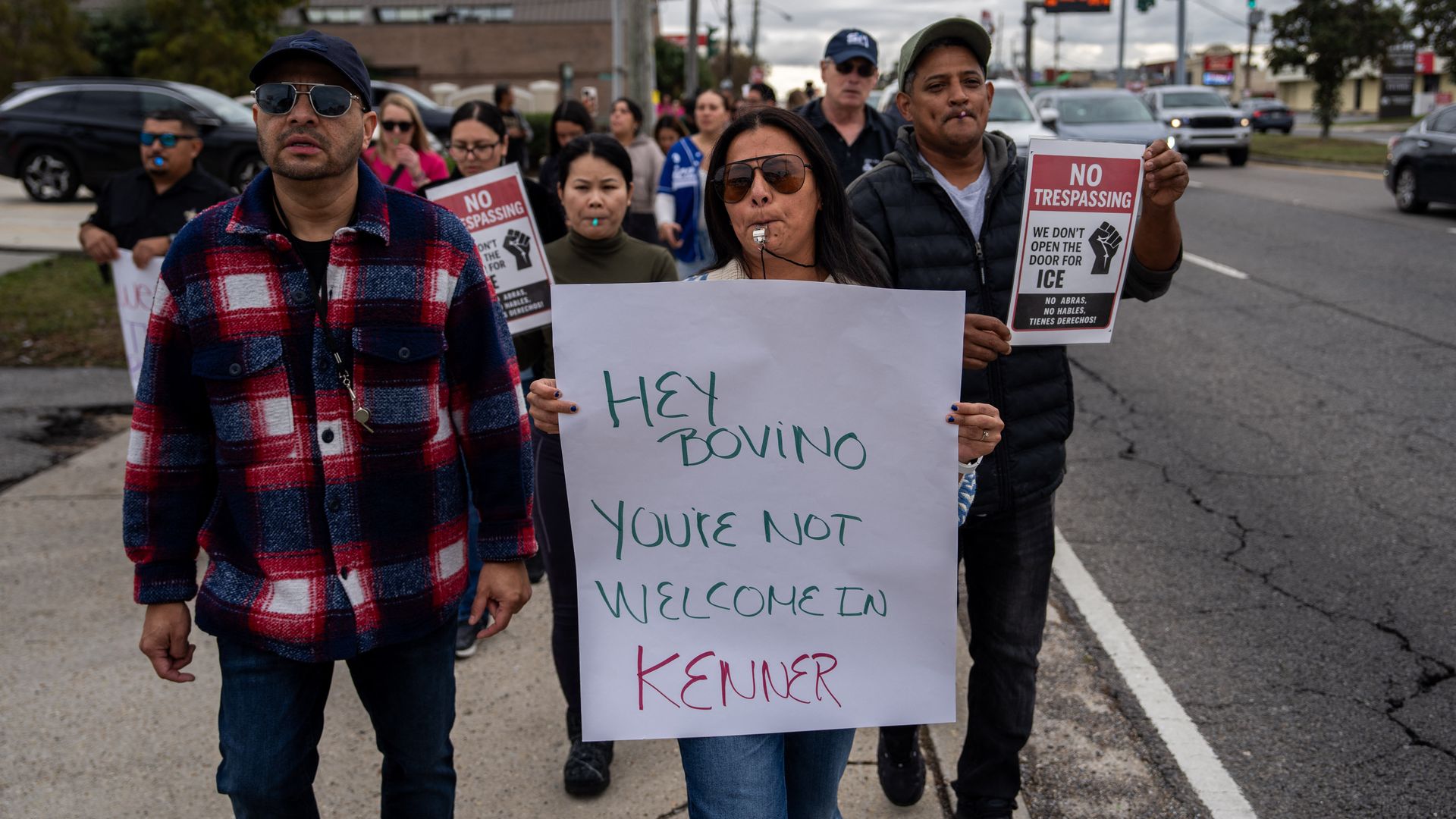 A crowd of people holding protest signs walks along a sidewalk next to a busy street of traffic. At the front, a woman blows on a whistle and holds a sign that says "Hey Bovino you're not welcome in Kenner."