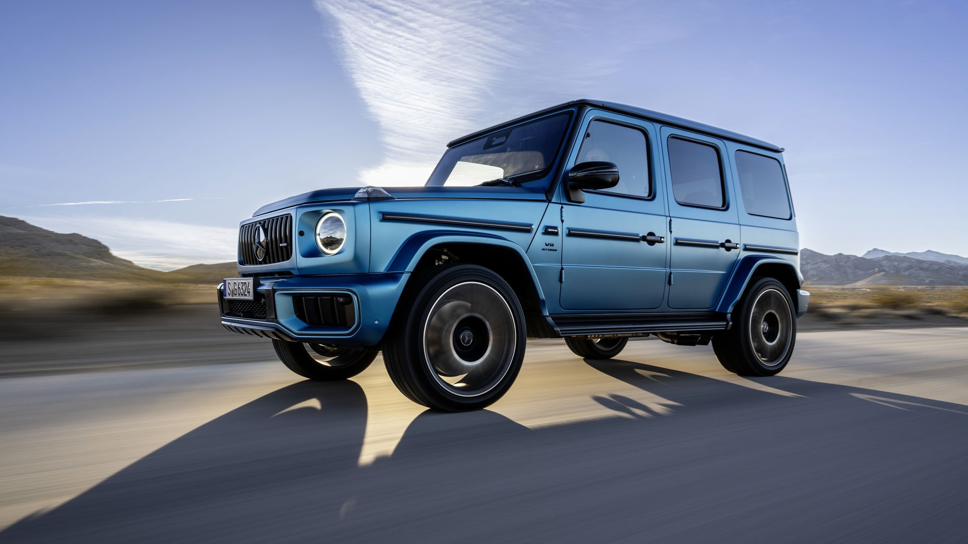 Blue Mercedes-Benz G-Class SUV driving on a desert road at sunset with mountains in the background and a partly cloudy sky.