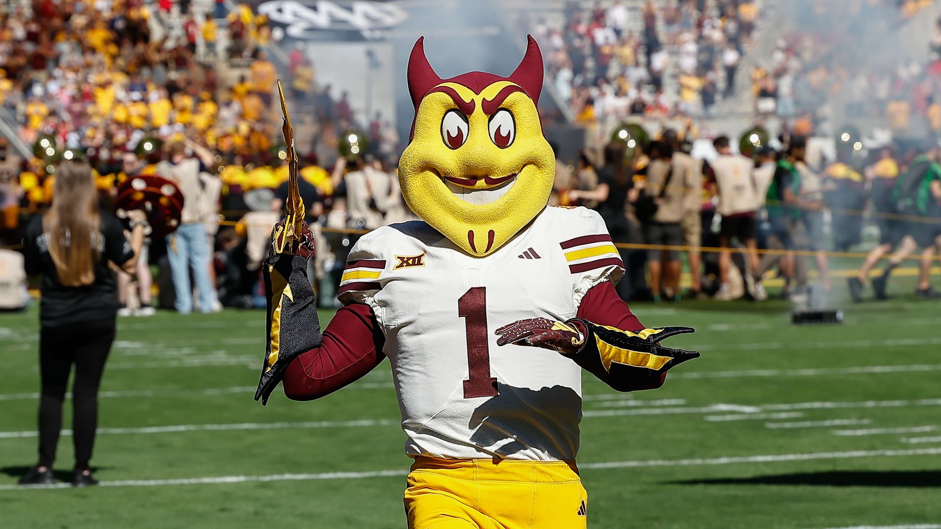 ASU's mascot Sun Devil walking on the field with crows on the bleachers in the background.