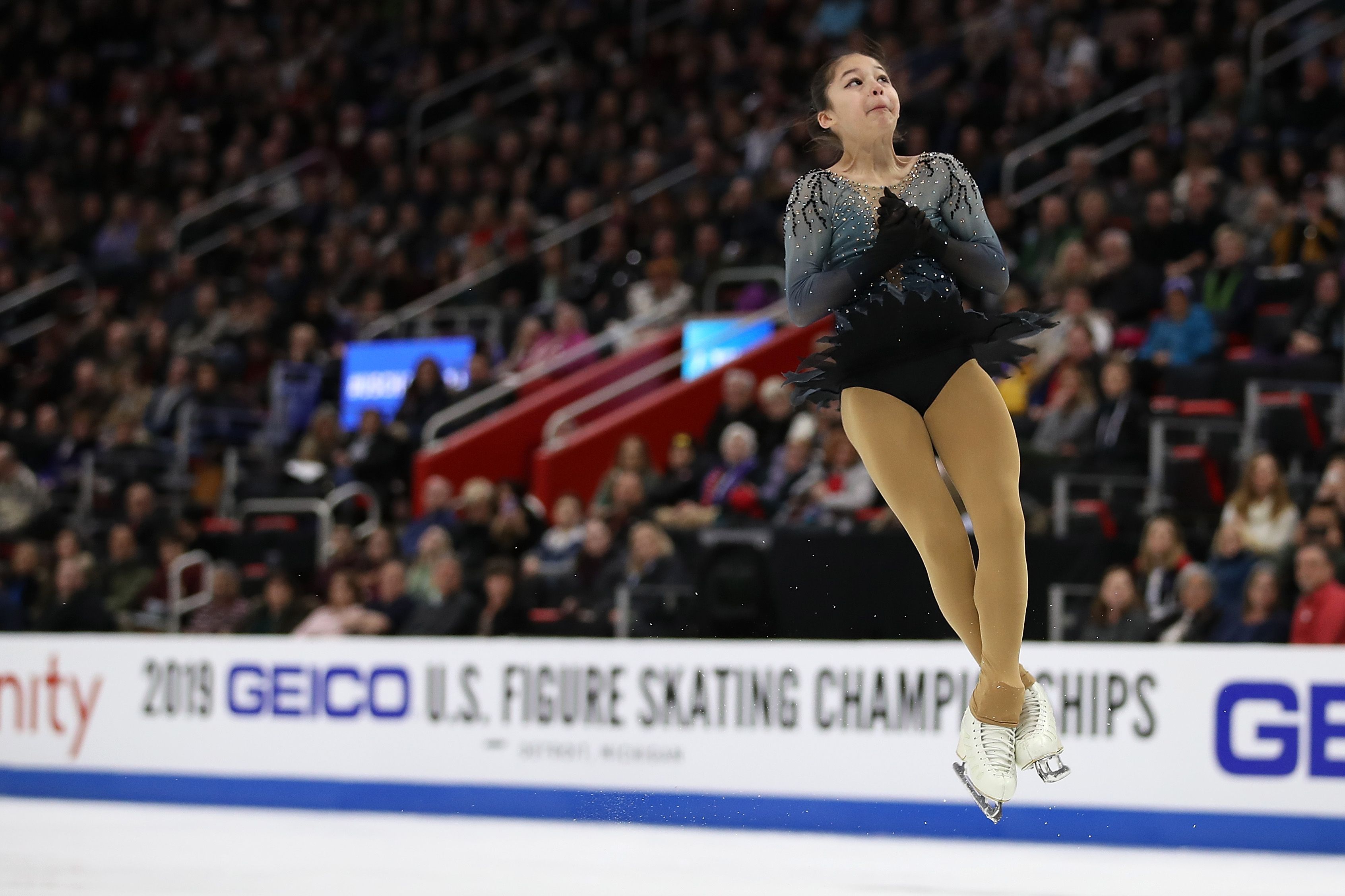 Figure skater in a sparkly black and gray costume mid-jump at the 2019 GEICO U.S. Figure Skating Championships, with a crowd watching in the background.
