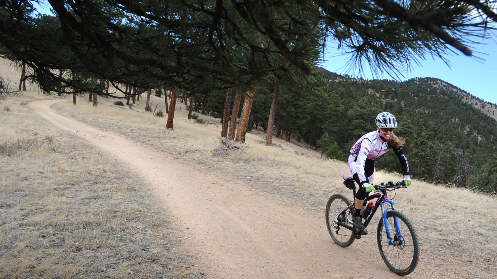Woman in a white helmet and purple cycling gear rides a mountain bike on a dirt path through a dry grassy area with pine trees and hills in the background under a clear sky.