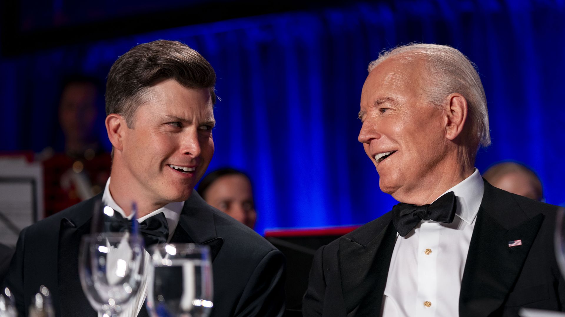 A photo showing the comedian Colin Jost on the left and former U.S. President Joe Biden on the right, both sitting at a table and wearing tuxedos.