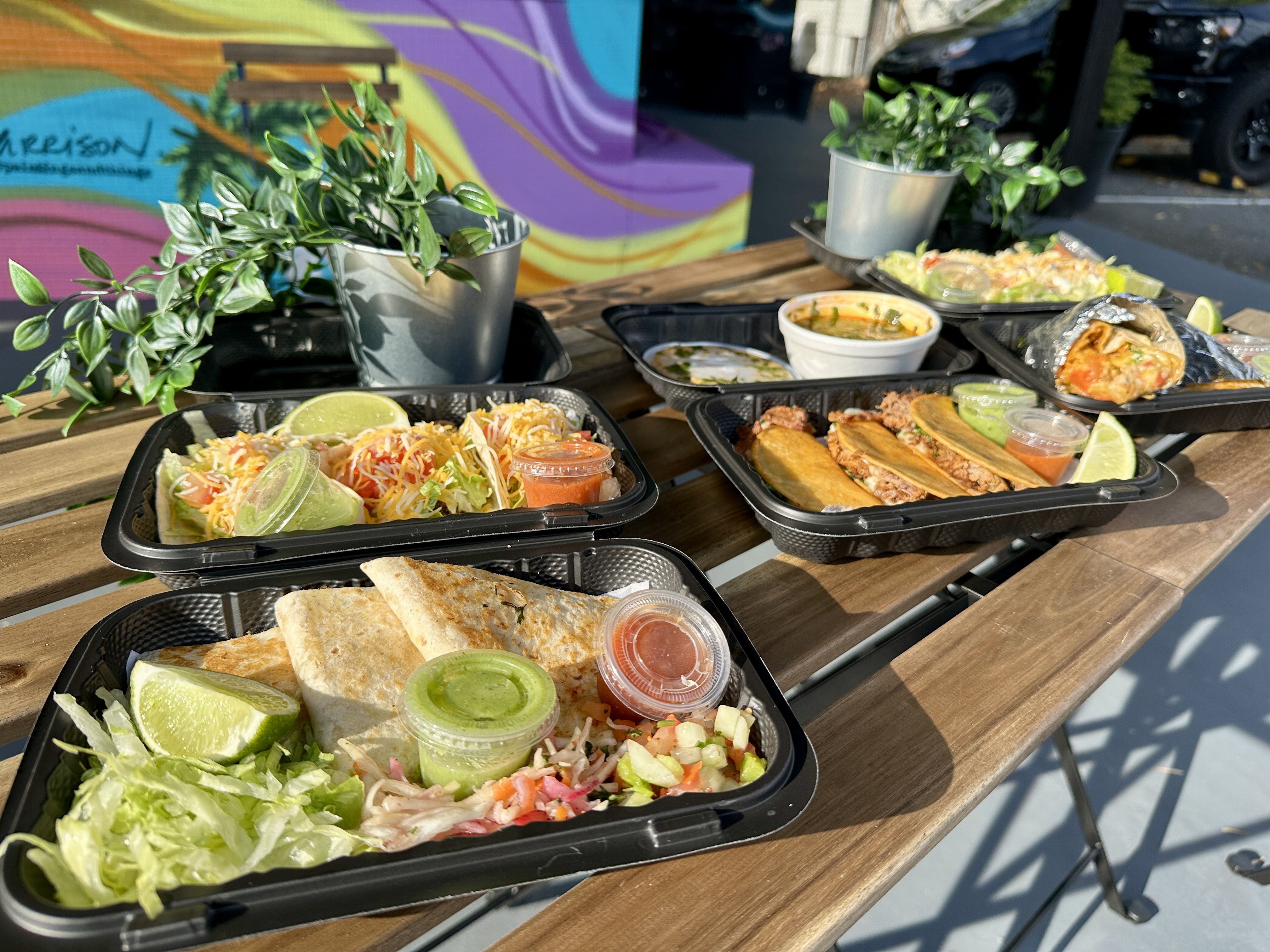 Outdoor wooden table with four black trays of Mexican food including tacos, quesadillas, salad, lime wedges, and sauces, with two small potted green plants and colorful mural background.
