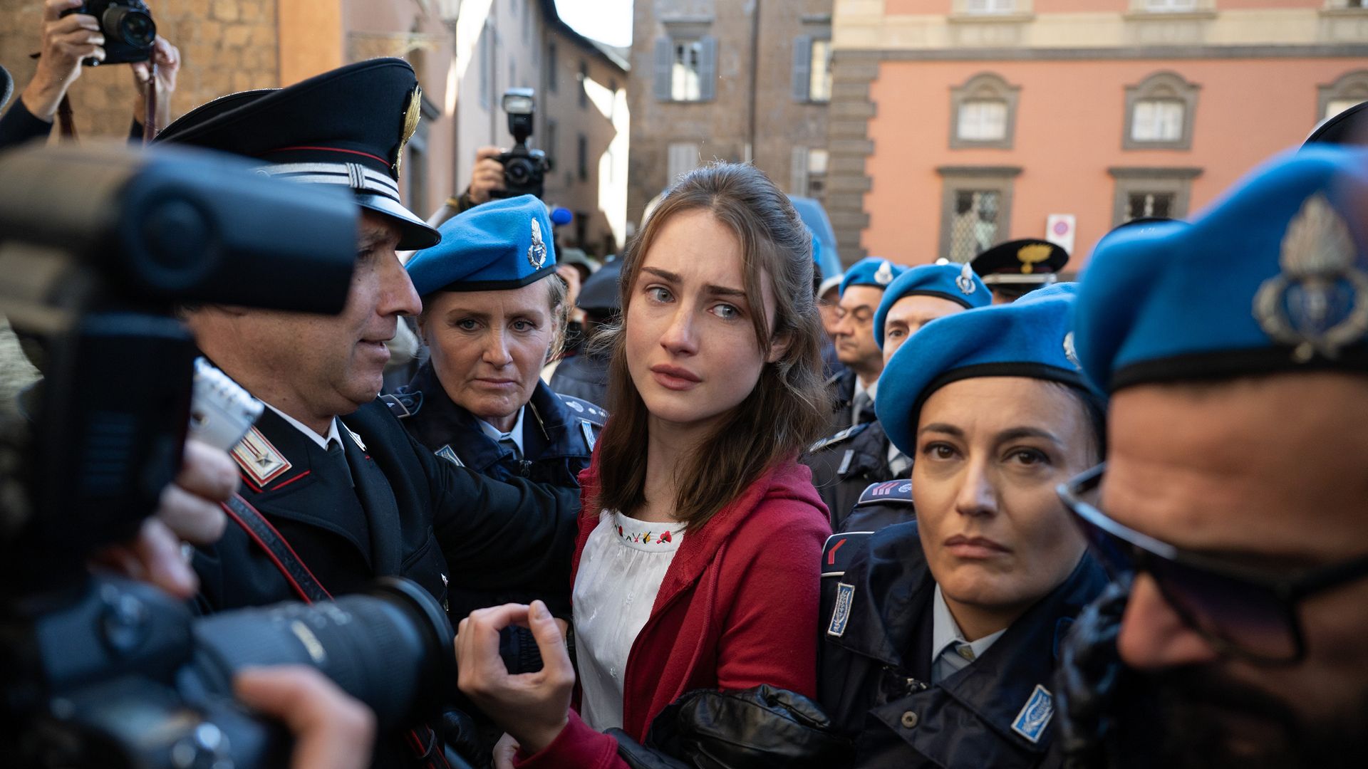 A woman with brown hair and a concerned look is surrounded by Italian police and media. 