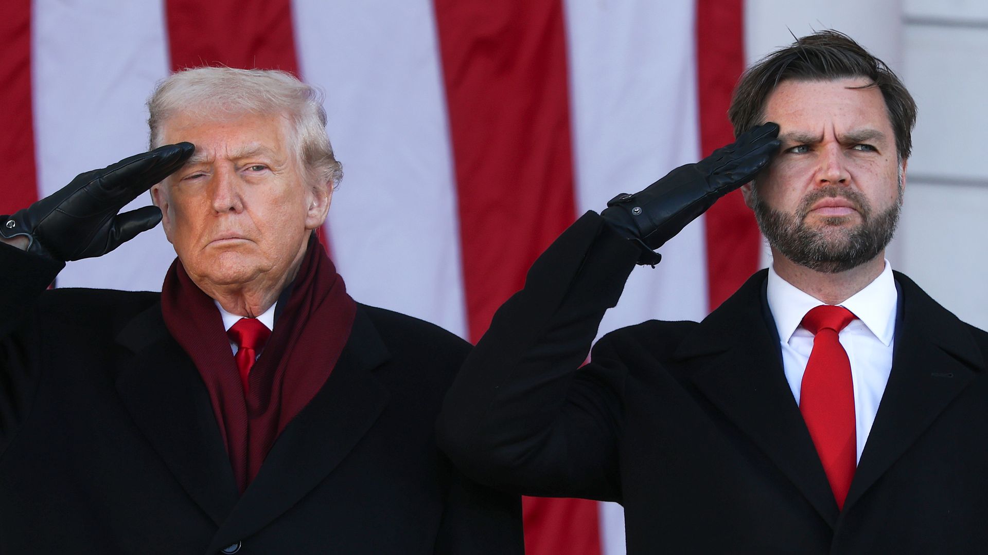 Two men in dark coats with red ties and black gloves salute with raised right hands, standing before a red-and-white striped backdrop, serious expressions.