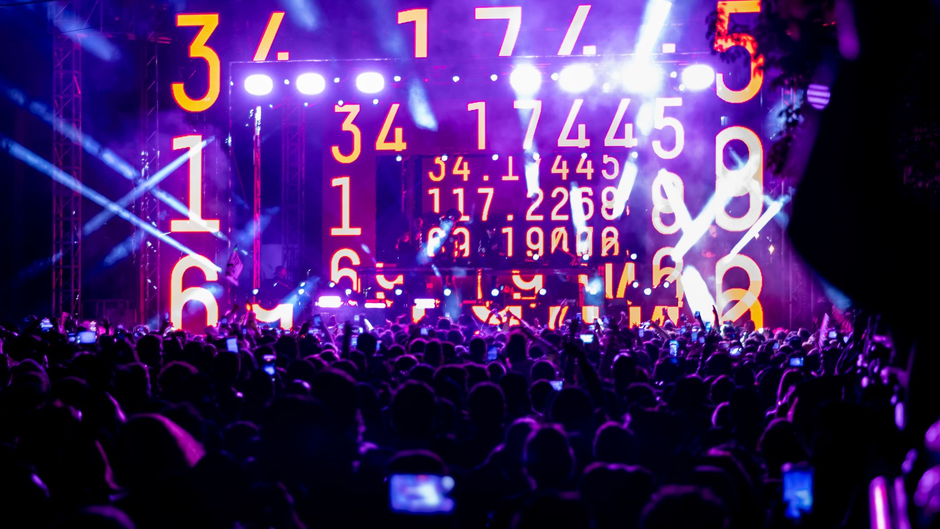 Photo of a crowd of people at an EDM concert featuring flashing lights and numbers projected onto the stage