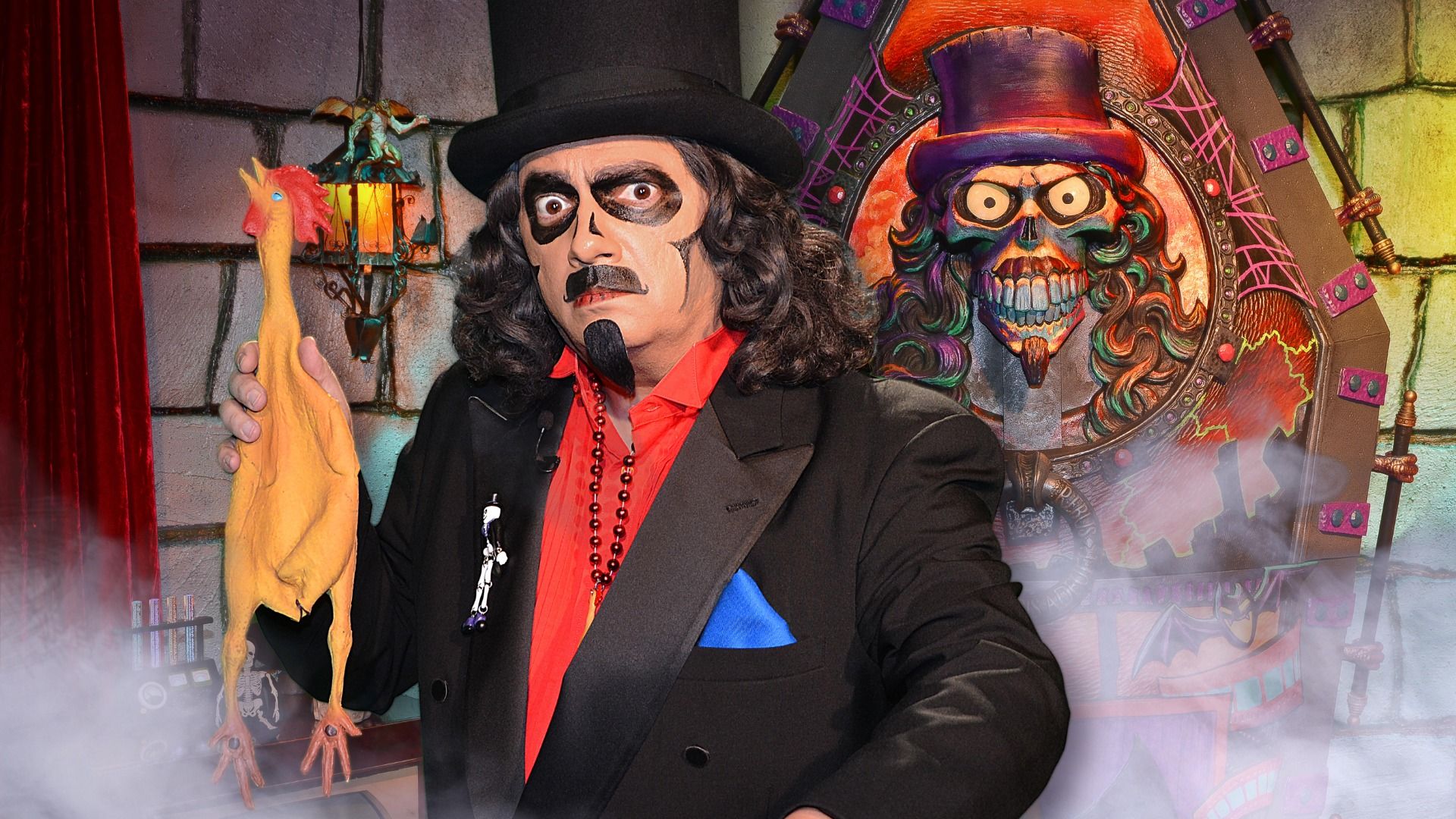 Man in skeleton face paint, black top hat and suit, red shirt, holding a rubber chicken. Colorful skull figure with purple hat in background, mist and spooky setting.