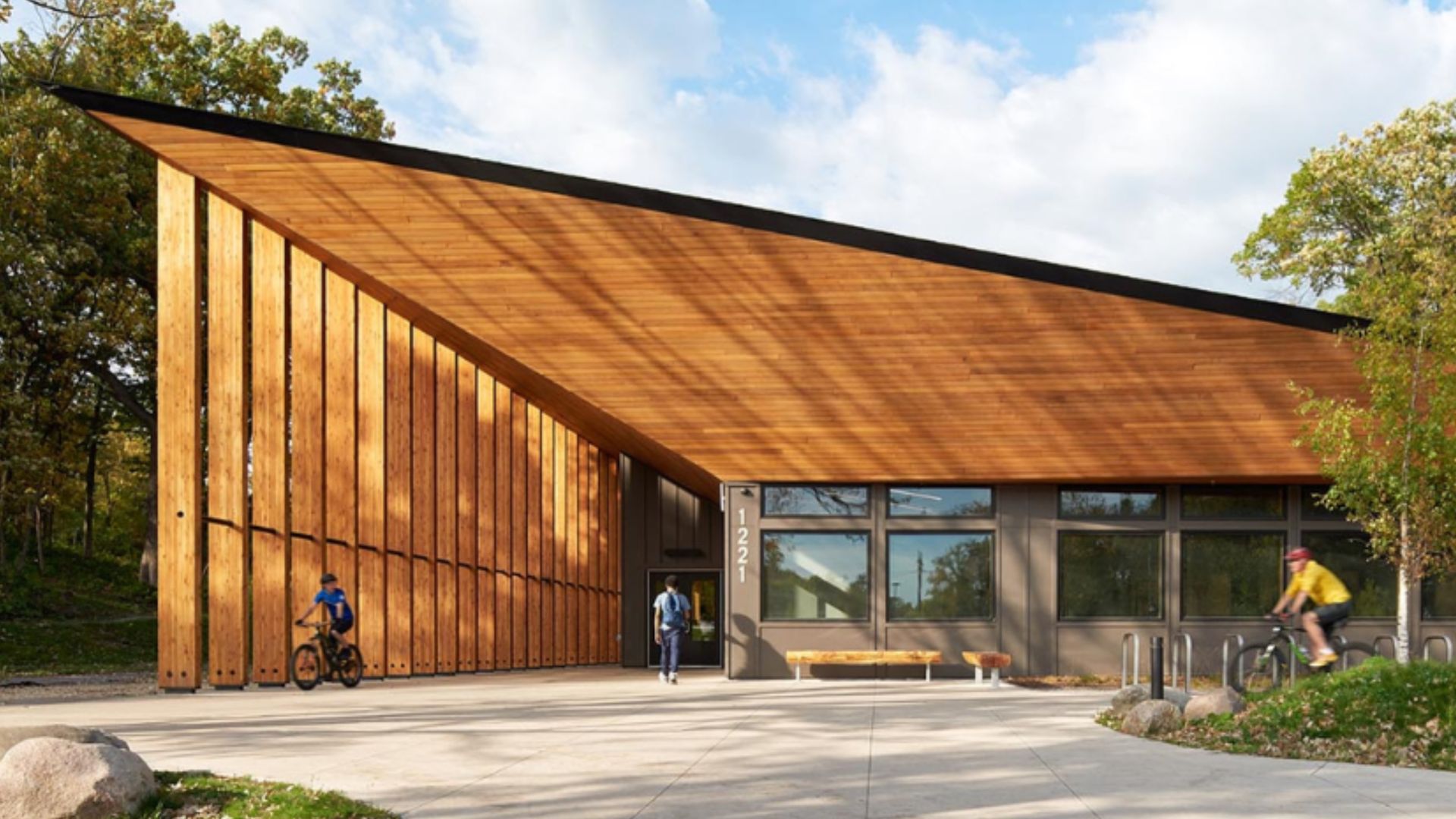 Modern wooden building with a long slanted copper roof and vertical slat wall on the left. Large front windows, trees, and blue sky. A person enters as cyclists ride by.