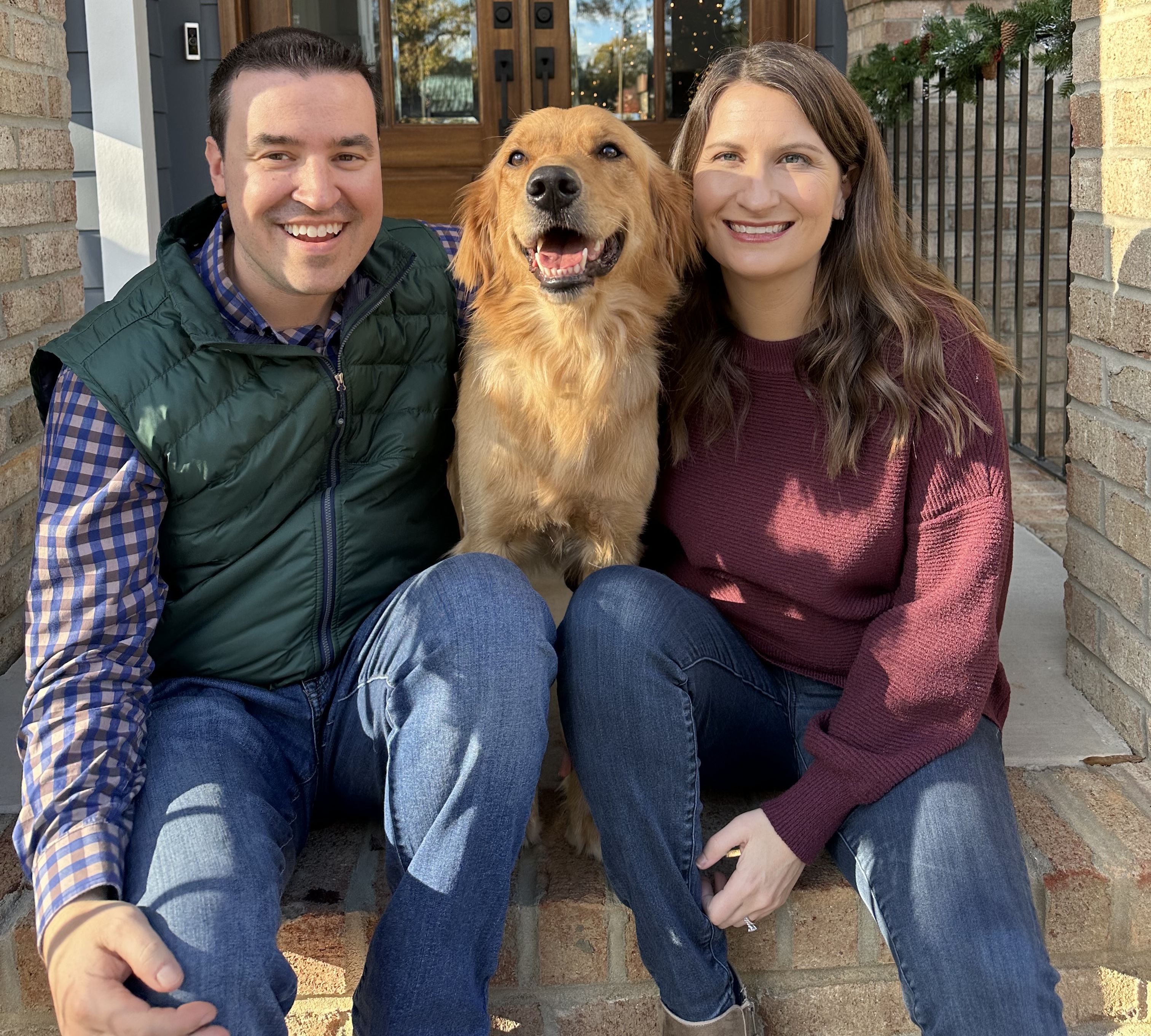 Smiling man in green vest and checkered shirt and woman in maroon sweater sitting on steps with golden retriever in between, in front of a door with holiday wreaths.