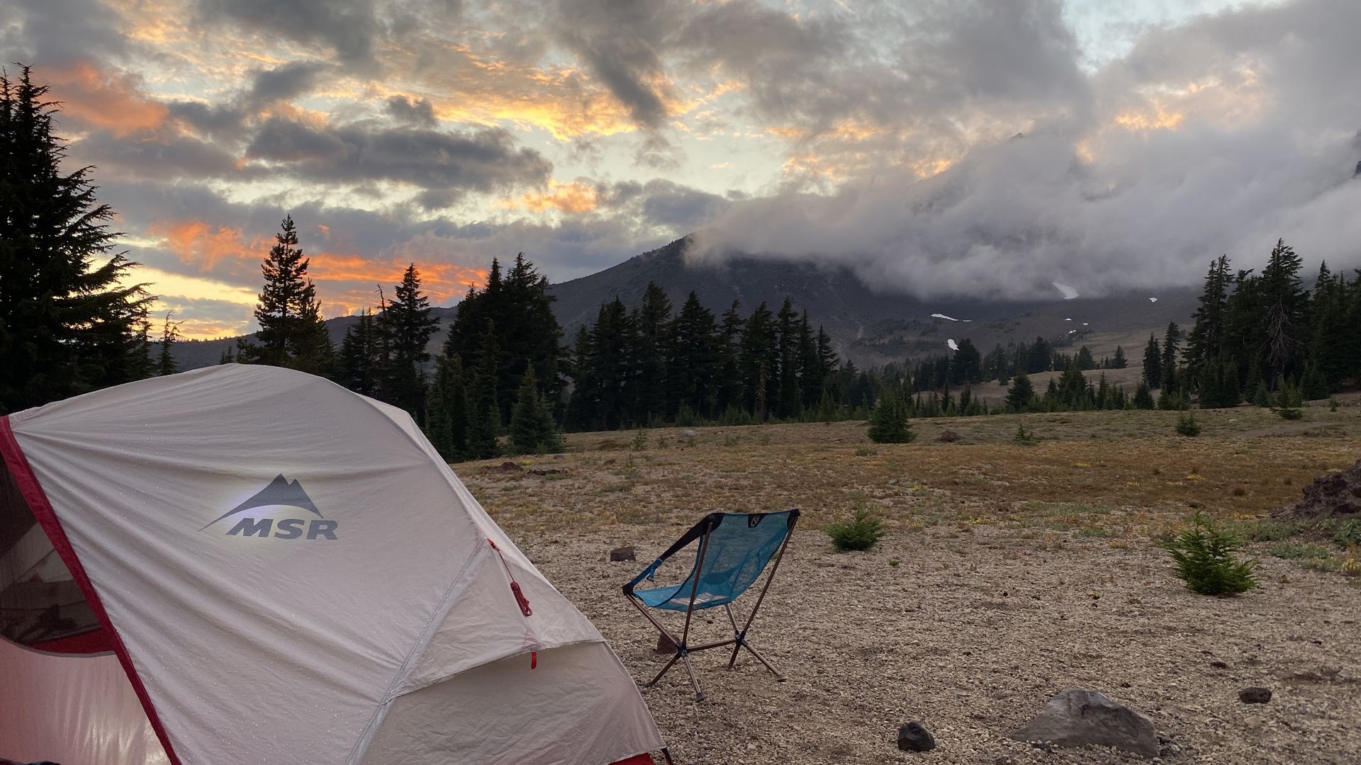 A tent and camping chair are seen in the foreground, with a colorful sunset over a mountain looms behind.