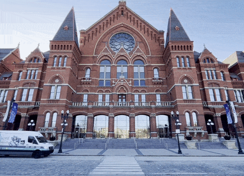 A large red-brick Gothic Revival building with a central rose window, arched windows, and two tall towers flanking the façade. Steps lead to glass doors; a white van sits to the left.