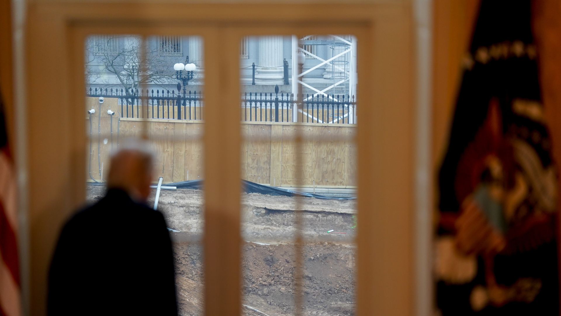 President Donald Trump stands inside the White House, looking toward construction work through a window.