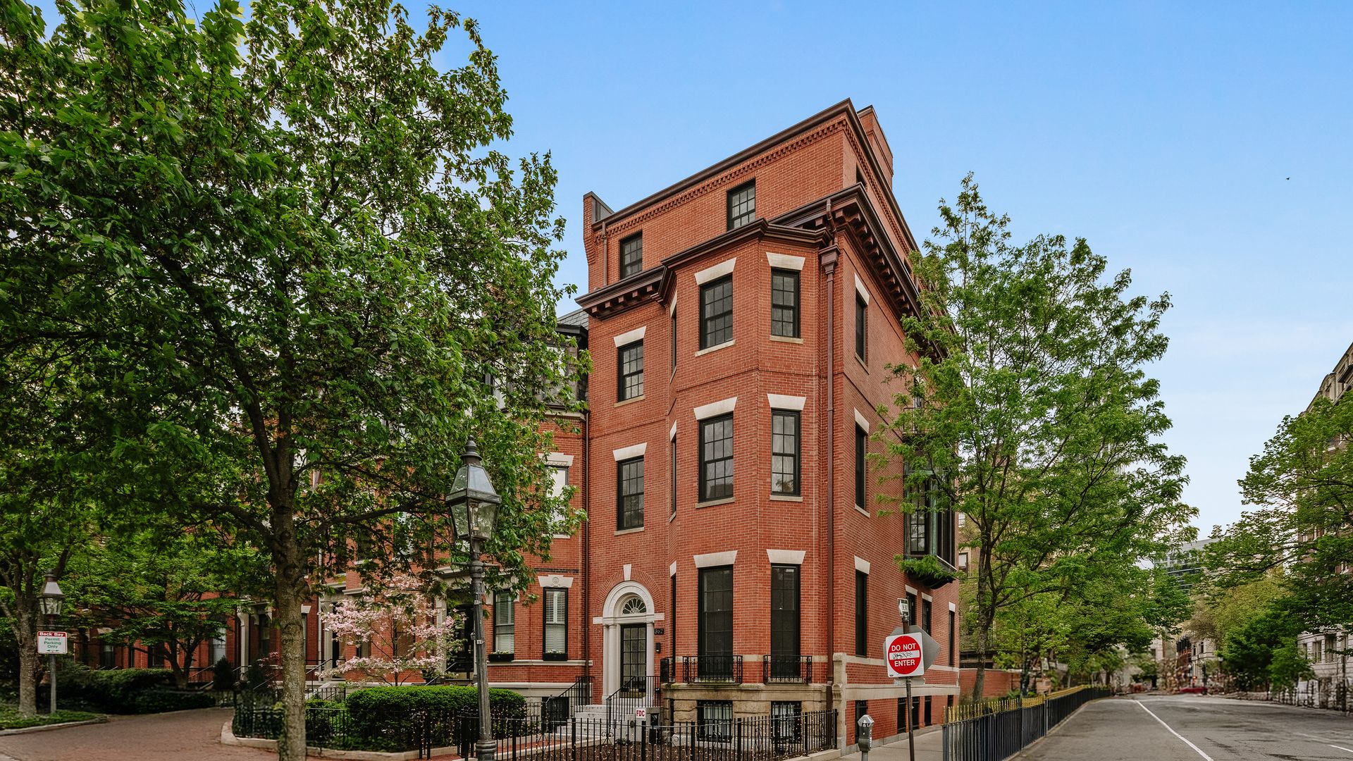 Red brick corner building with black windows and white trim, surrounded by green trees and a brick sidewalk with a vintage-style street lamp and a "Do Not Enter" sign.