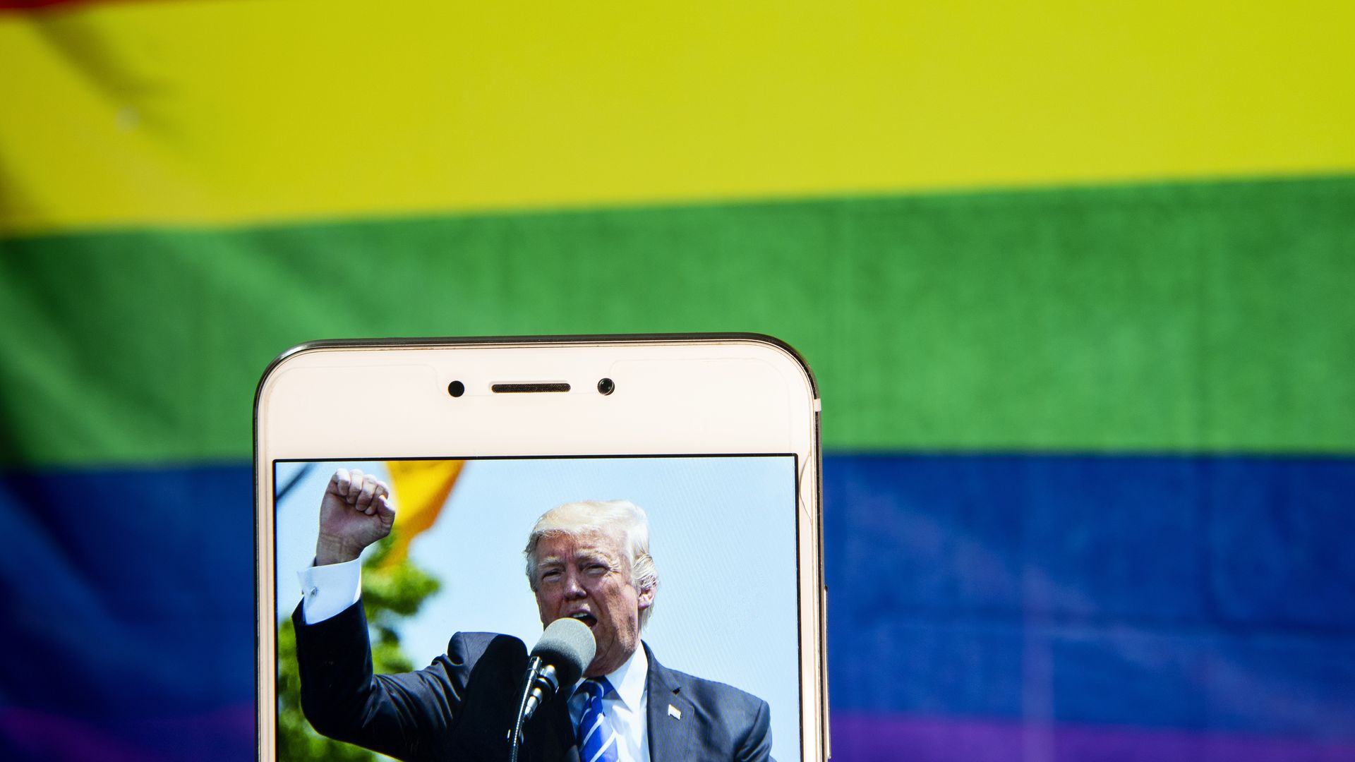 In this image, a picture of Donald Trump on a phone is held in front of a rainbow pride flag.