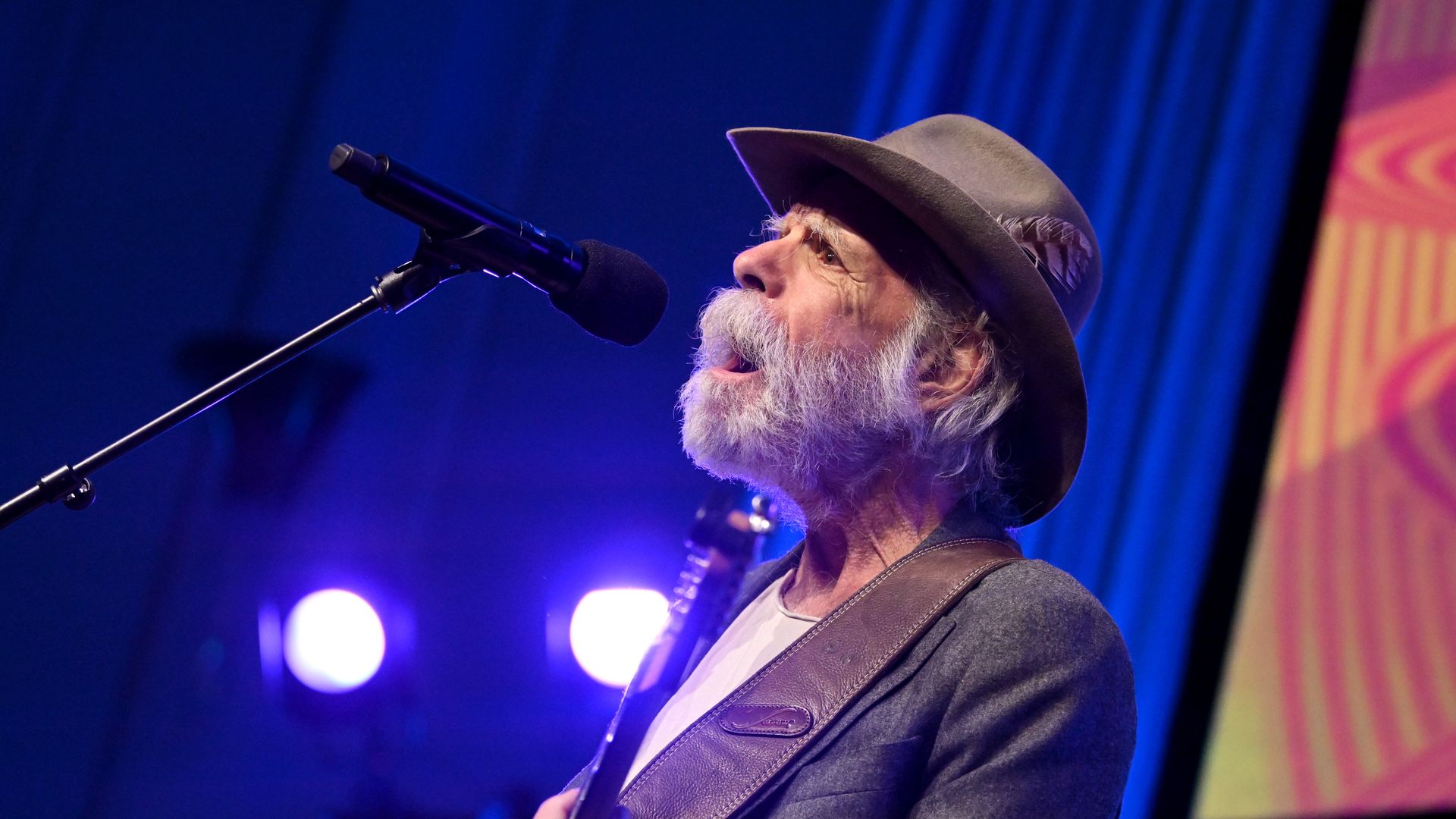 Elderly man with white beard and mustache wearing a hat and gray jacket singing into microphone on stage with blue lights and colorful backdrop.