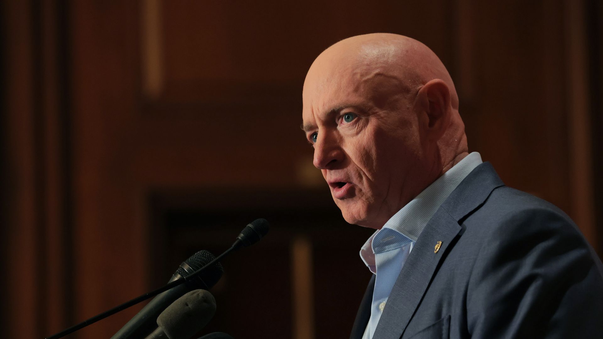 Kelly wearing a dark suit and slightly unbuttoned light blue shirt speaking into a mic