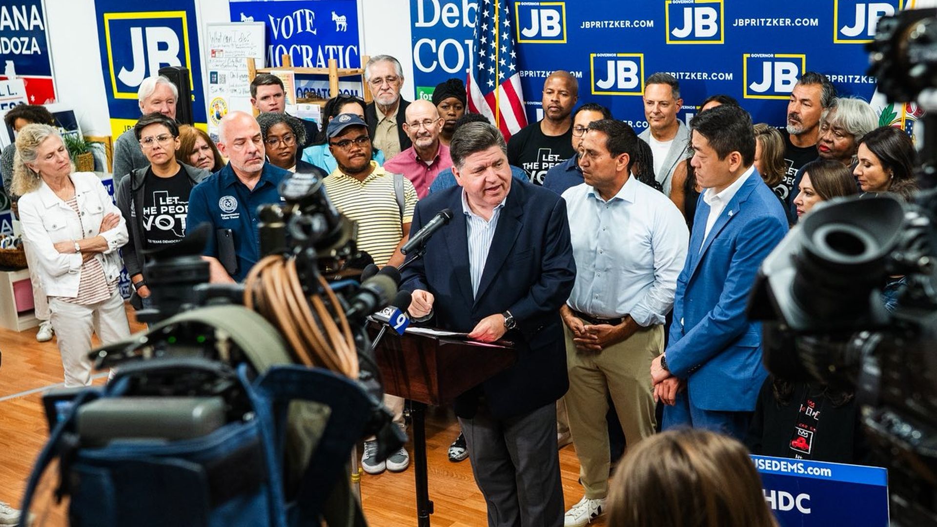 A political press conference with Illinois Gov. JB Pritzker speaking at a podium surrounded by Texas Democrats, cameras, and signs supporting JB Pritzker and Democratic candidates in a room with a U.S. flag.