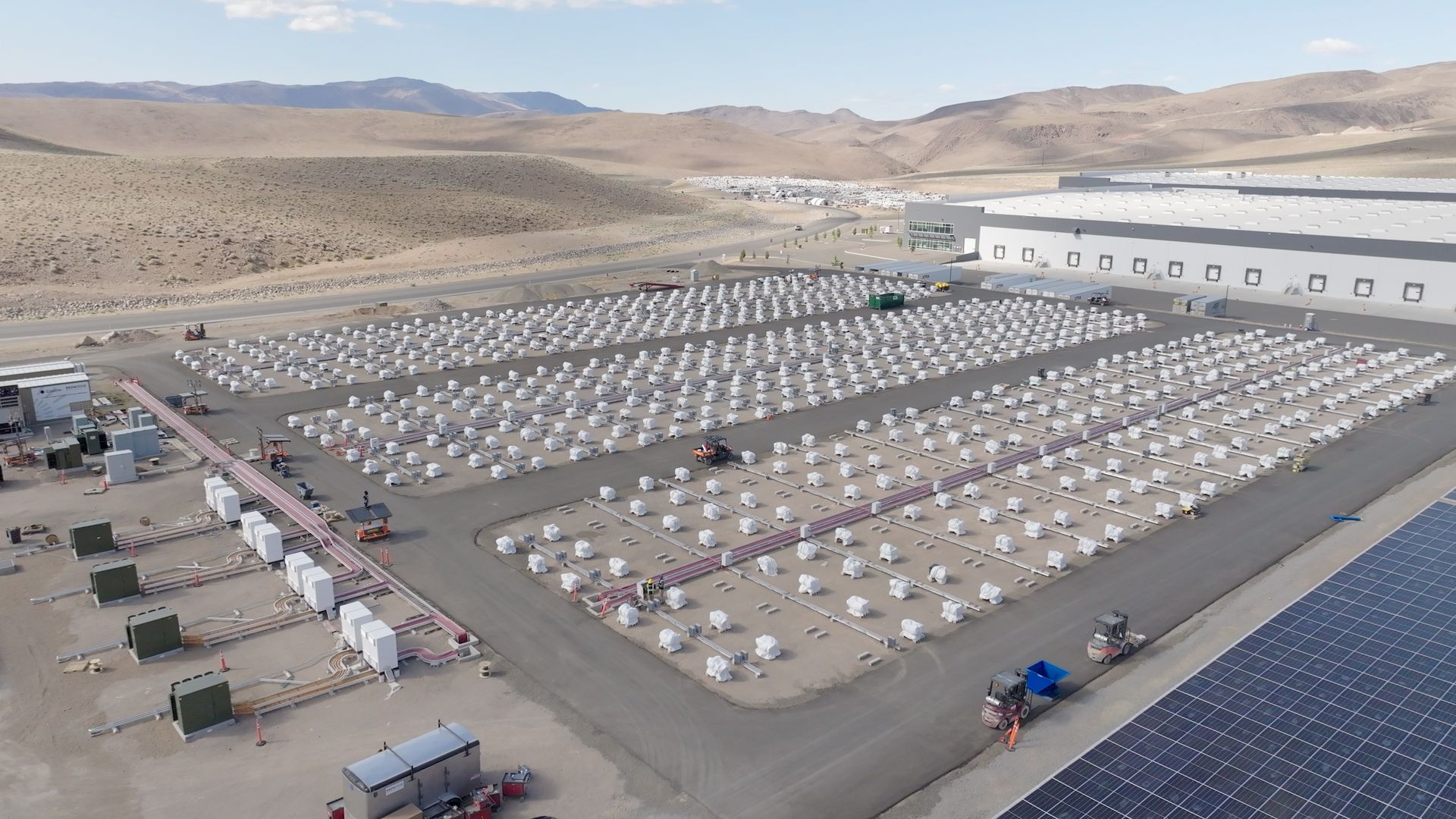 A large industrial site in a desert area with hundreds of white covered units arranged in rows, a gray building, and solar panels in the foreground under a clear blue sky.